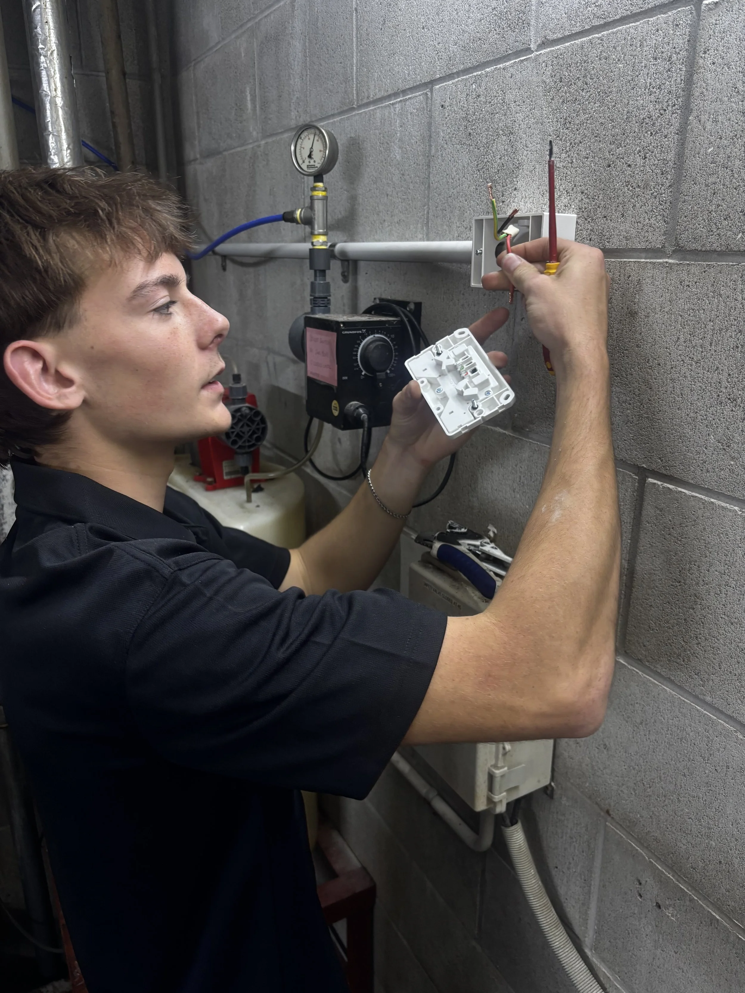 A technician working on electrical wiring inside an electrical box on a gray cinder block wall.