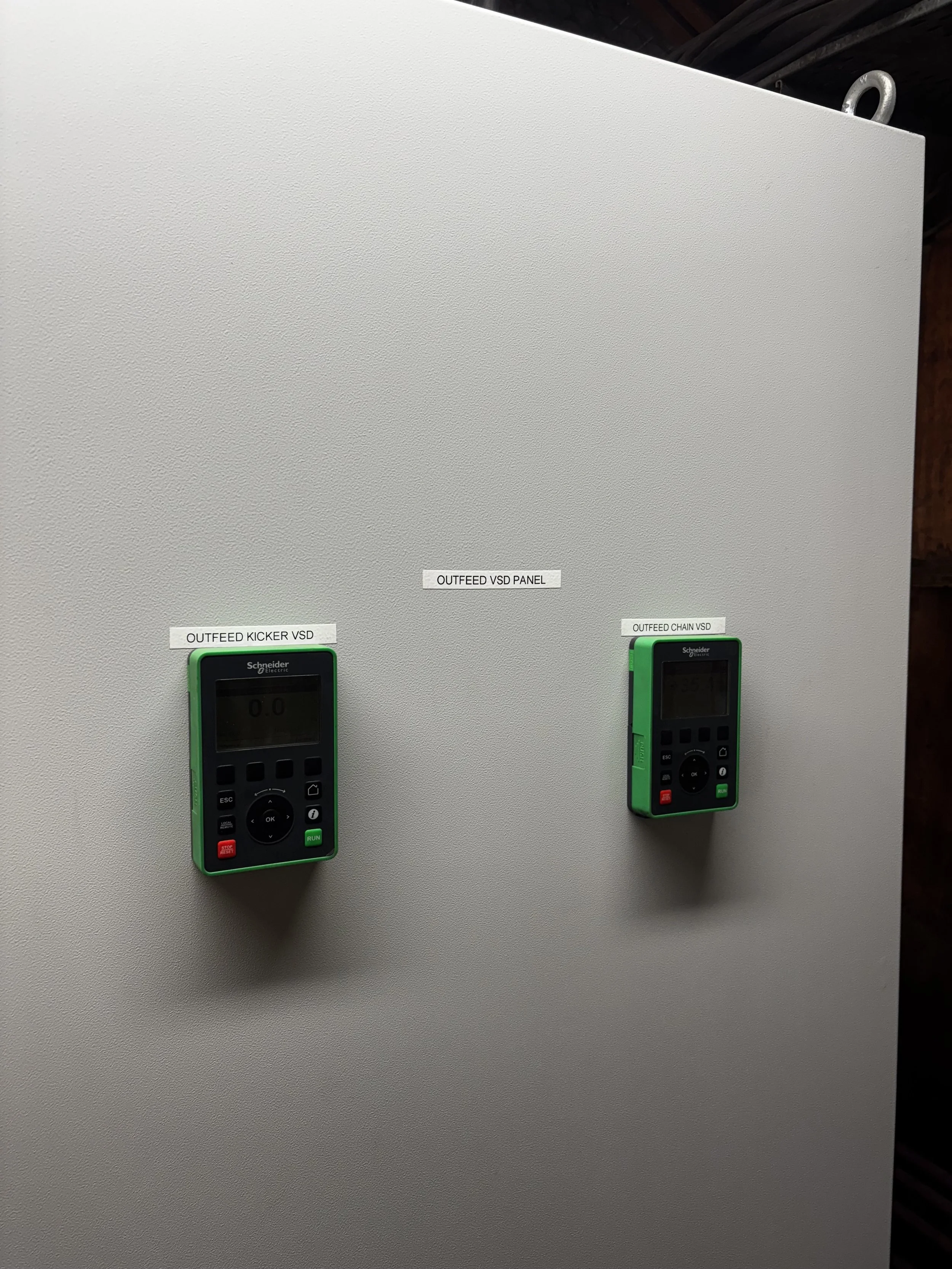 Electrical panel with two green Schneider controllers labeled 'OUTFEED KICKER VSD' and 'OUTFEED CHAIN VSD' mounted on a plain grey surface.