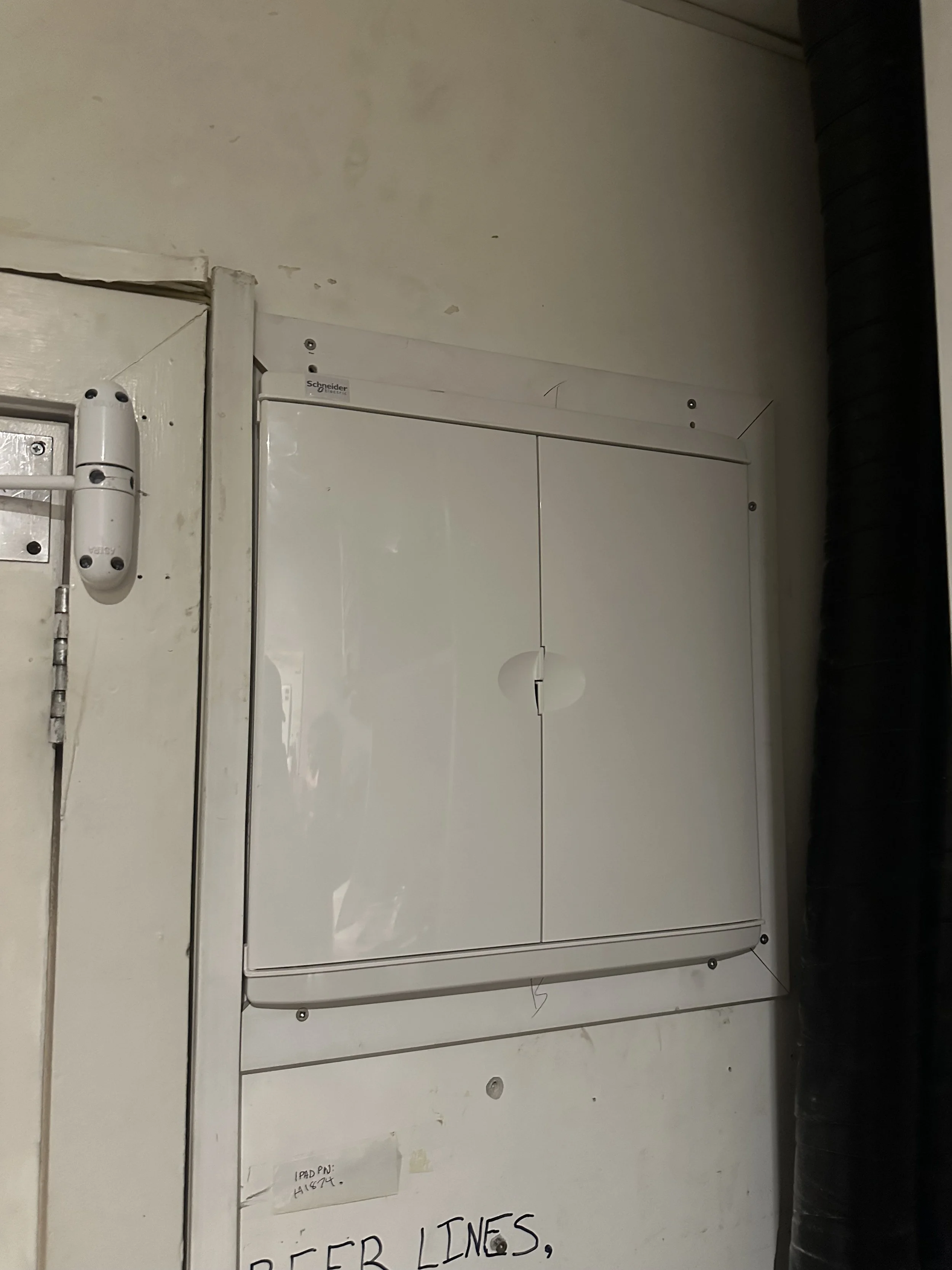 A white electrical or utility panel mounted on a wall with handwritten notes and a black flexible conduit on the right.