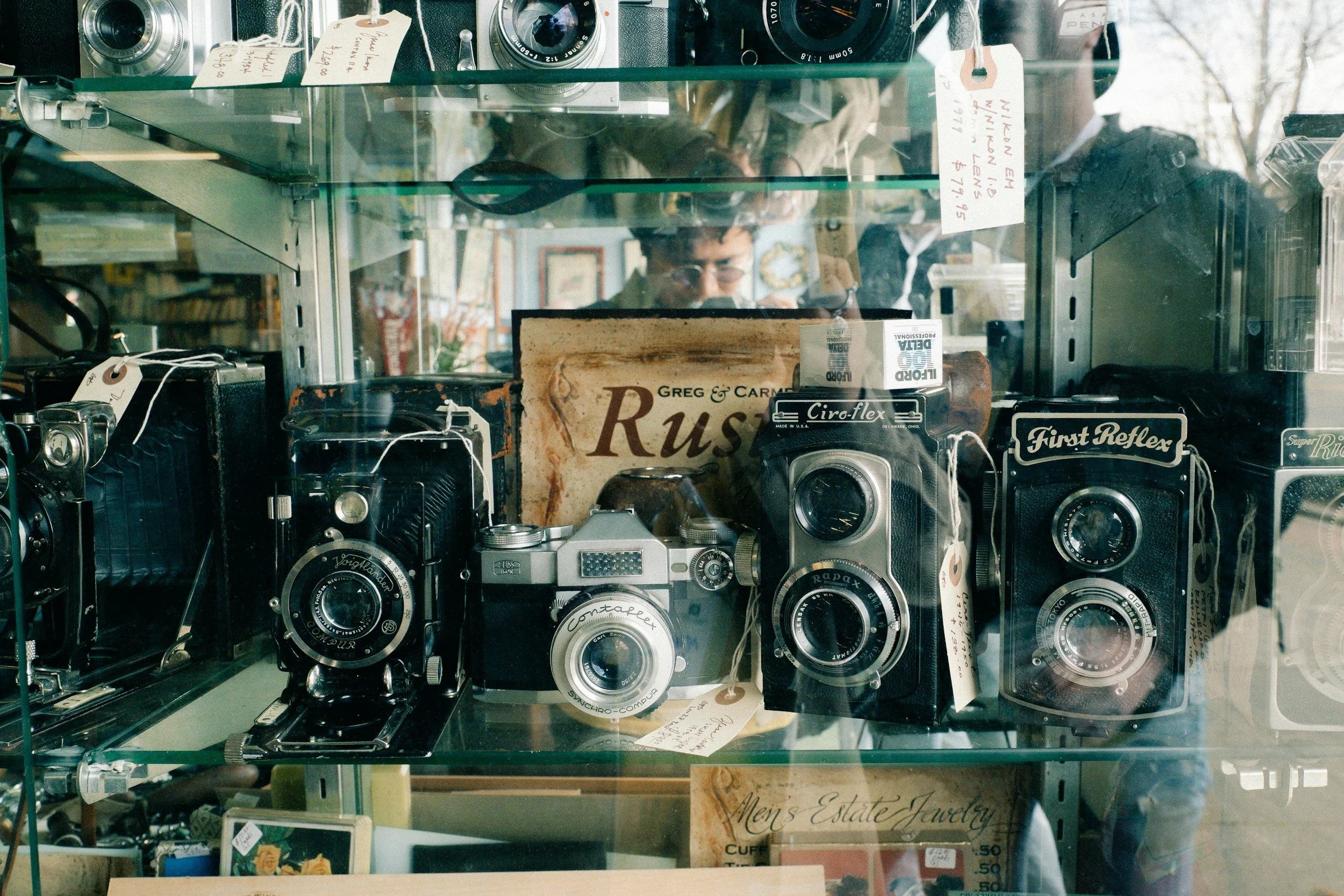 A glass display case filled with vintage cameras, including models from the 1950s and 1960s, with handwritten price tags and labels, and a person reflecting in the glass in the background.