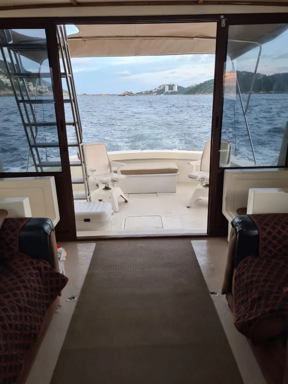 View of water and distant hills from the interior of a boat, with a sliding glass door opening onto the deck with seats and a ladder.