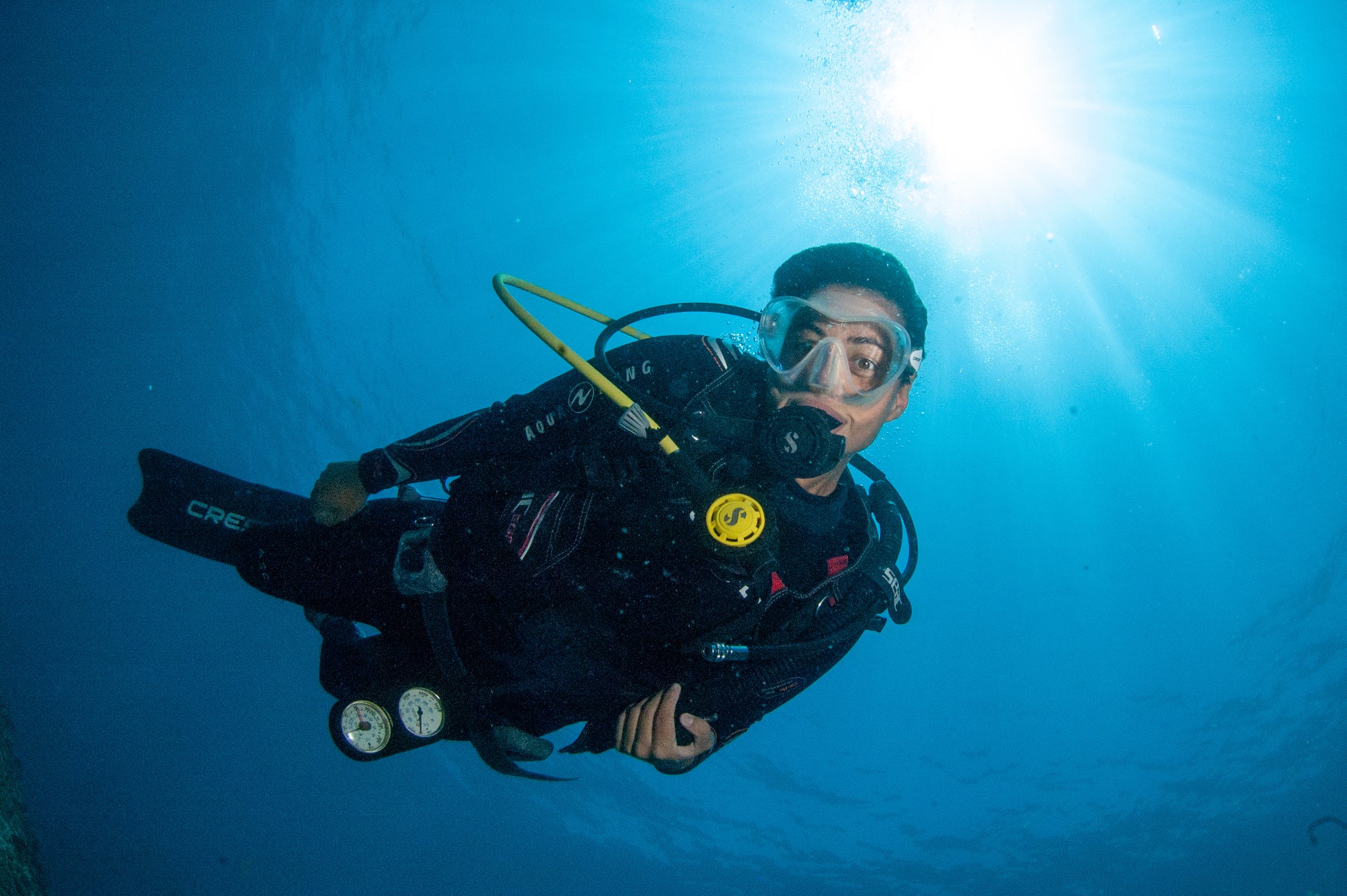 A man scuba diving underwater with sunlight shining from above, wearing a wetsuit, mask, and breathing apparatus.
