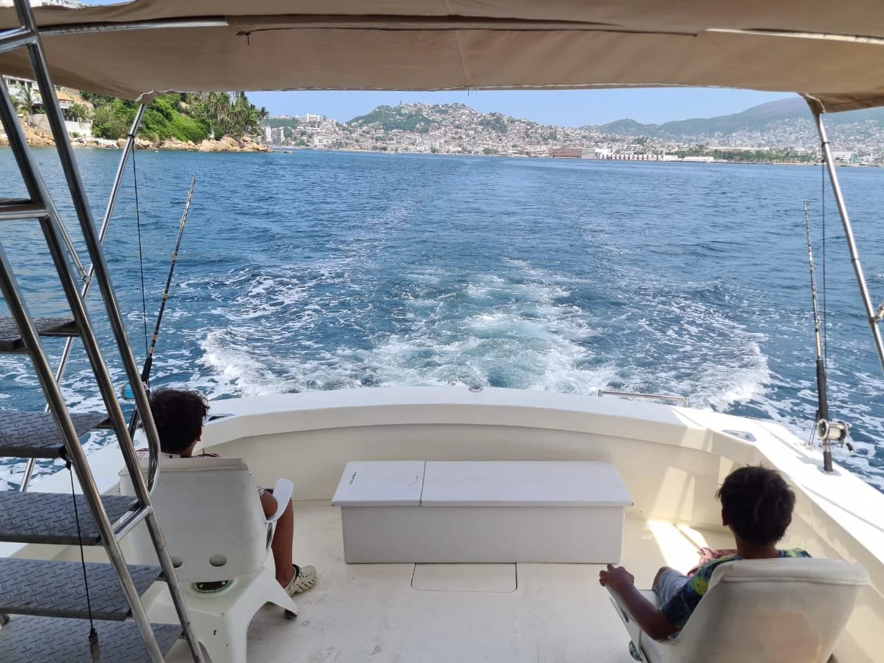 Two children sitting on a boat, looking at the water and distant shoreline with buildings and hills.