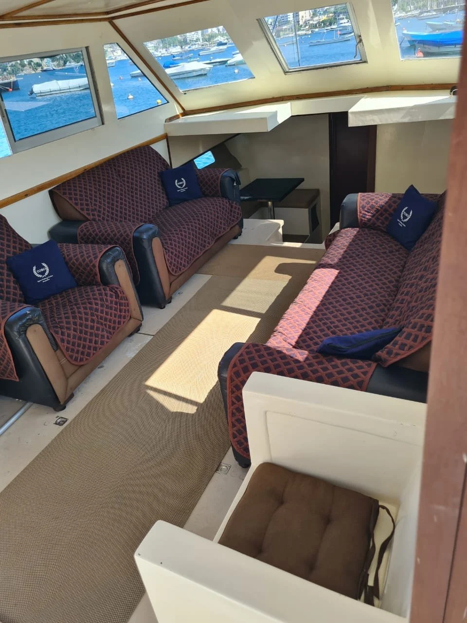Interior of a boat's lounge area with two benches and one armchair, featuring patterned cushions and windows showing a marina with boats.