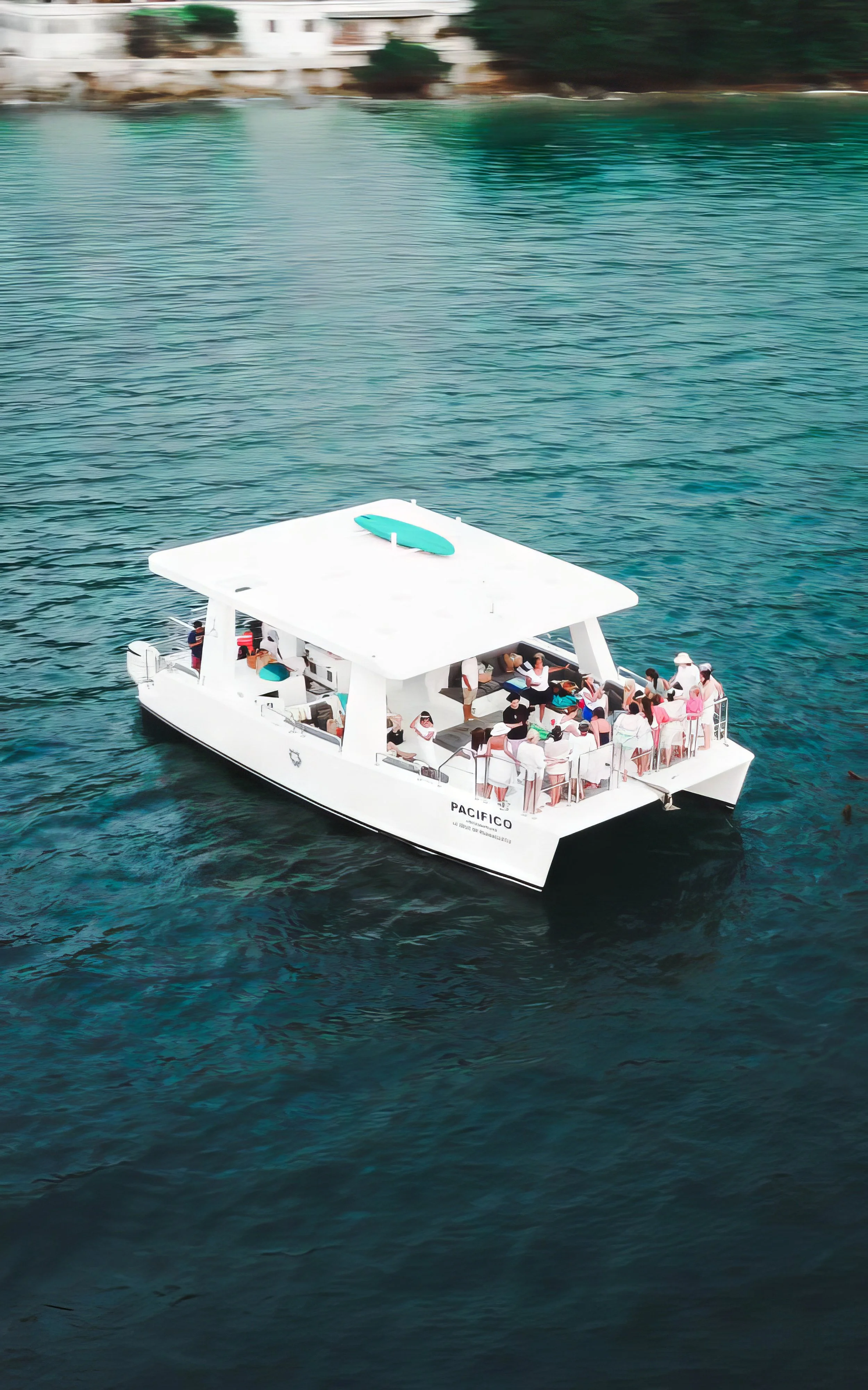 A white passenger boat named Pacifico floating on deep blue water with groups of people on board and a small surfboard on the roof.