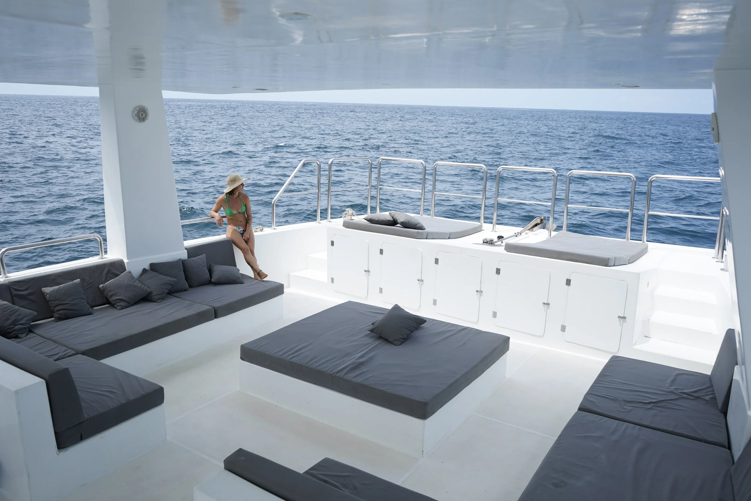 Woman in a green bikini and straw hat sitting on the edge of a white yacht's deck, looking out over the open ocean with blue water and sky.