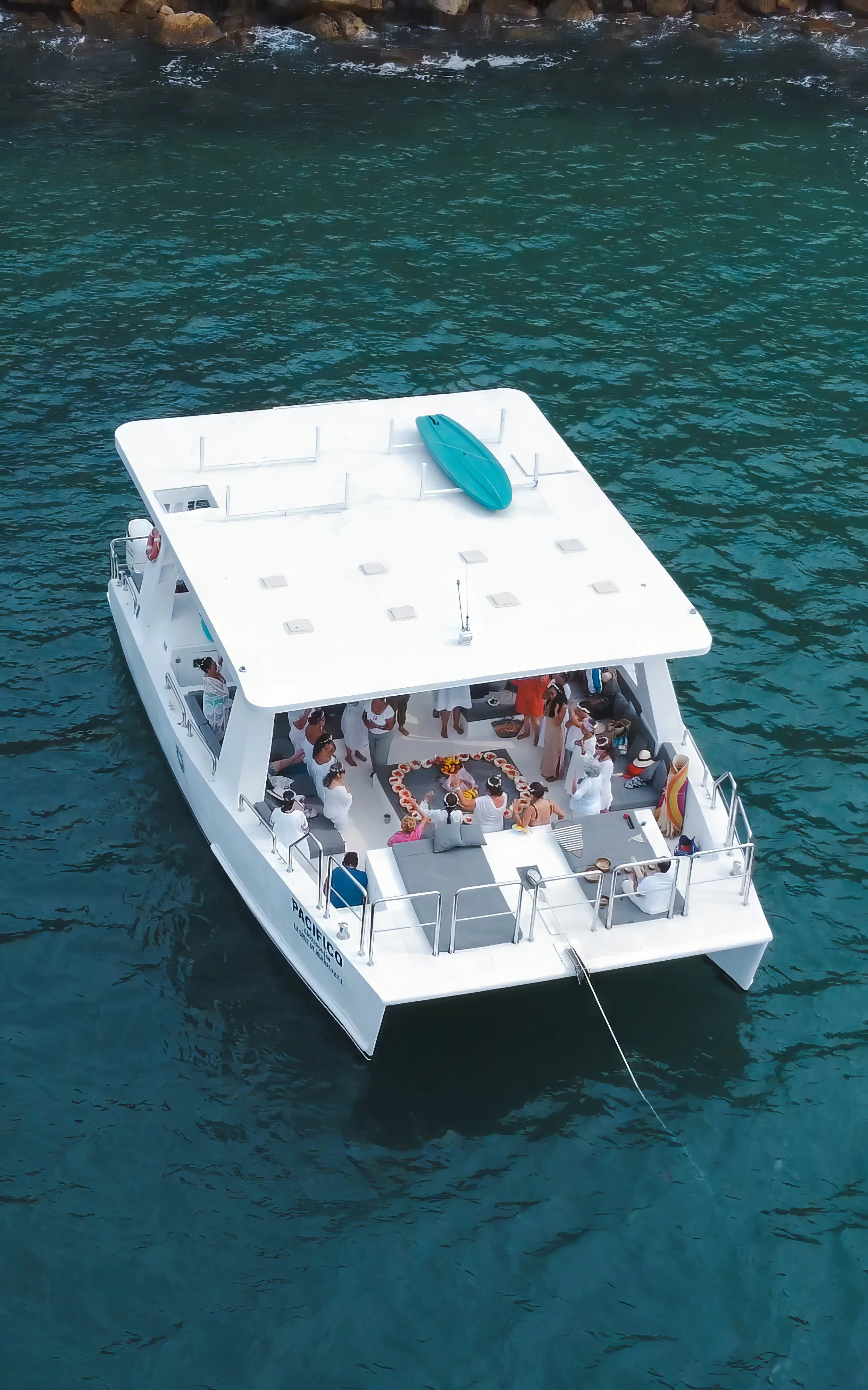 A group of people gathered on a white yacht celebrating around a cake, with some standing and some sitting, in clear blue water near rocky shoreline, with a paddleboard on the roof.
