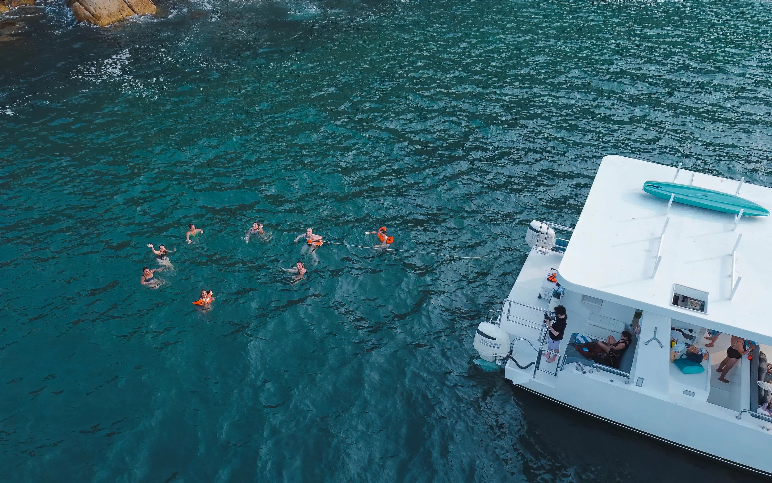 People swimming in clear blue water near a white boat with a teal surfboard on top