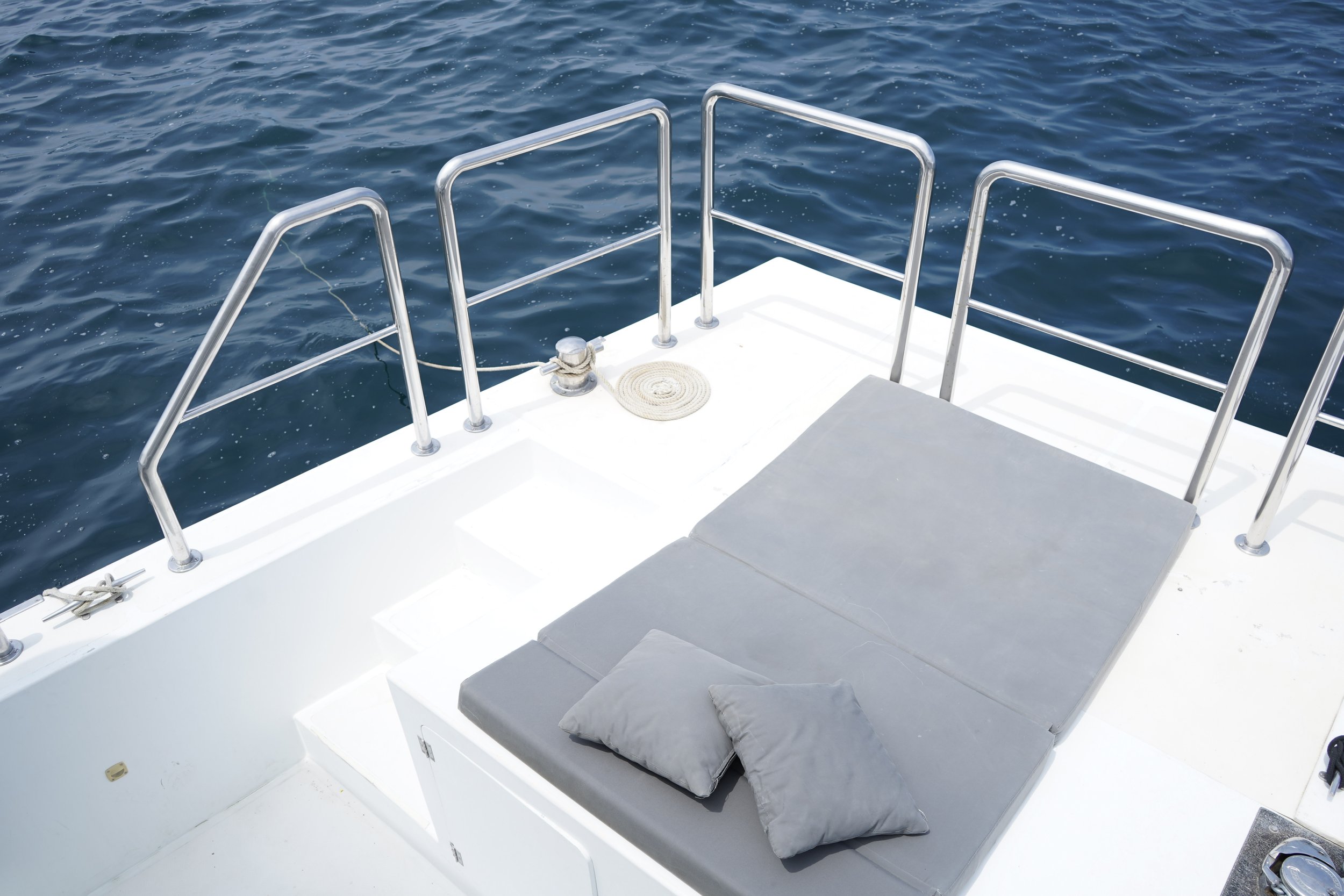 View of a boat deck with gray cushions and pillows, stainless steel railing, and blue water surrounding it.