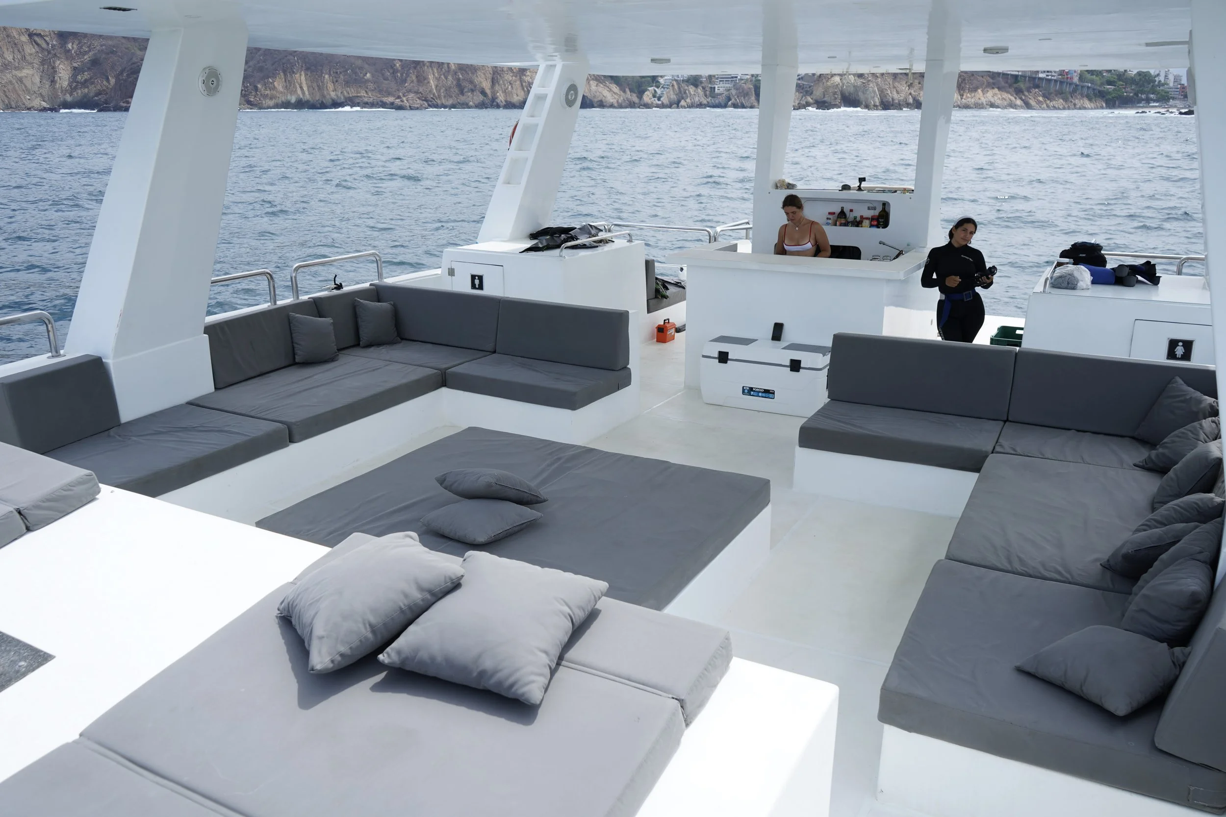 View of a boat deck with gray cushioned seating, pillows, and two women working behind the bar area, with ocean and cliffs in the background.