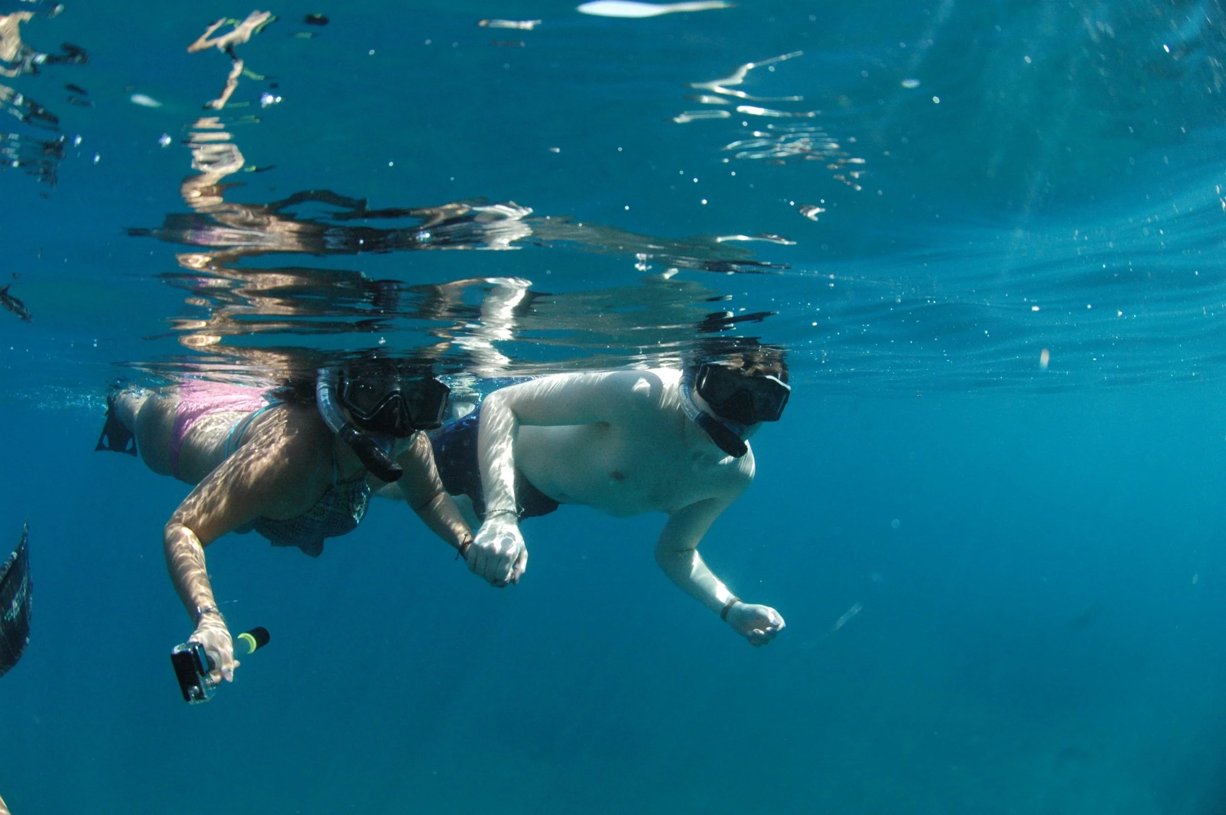 A woman and a man snorkeling underwater, holding hands, with a camera in her other hand.
