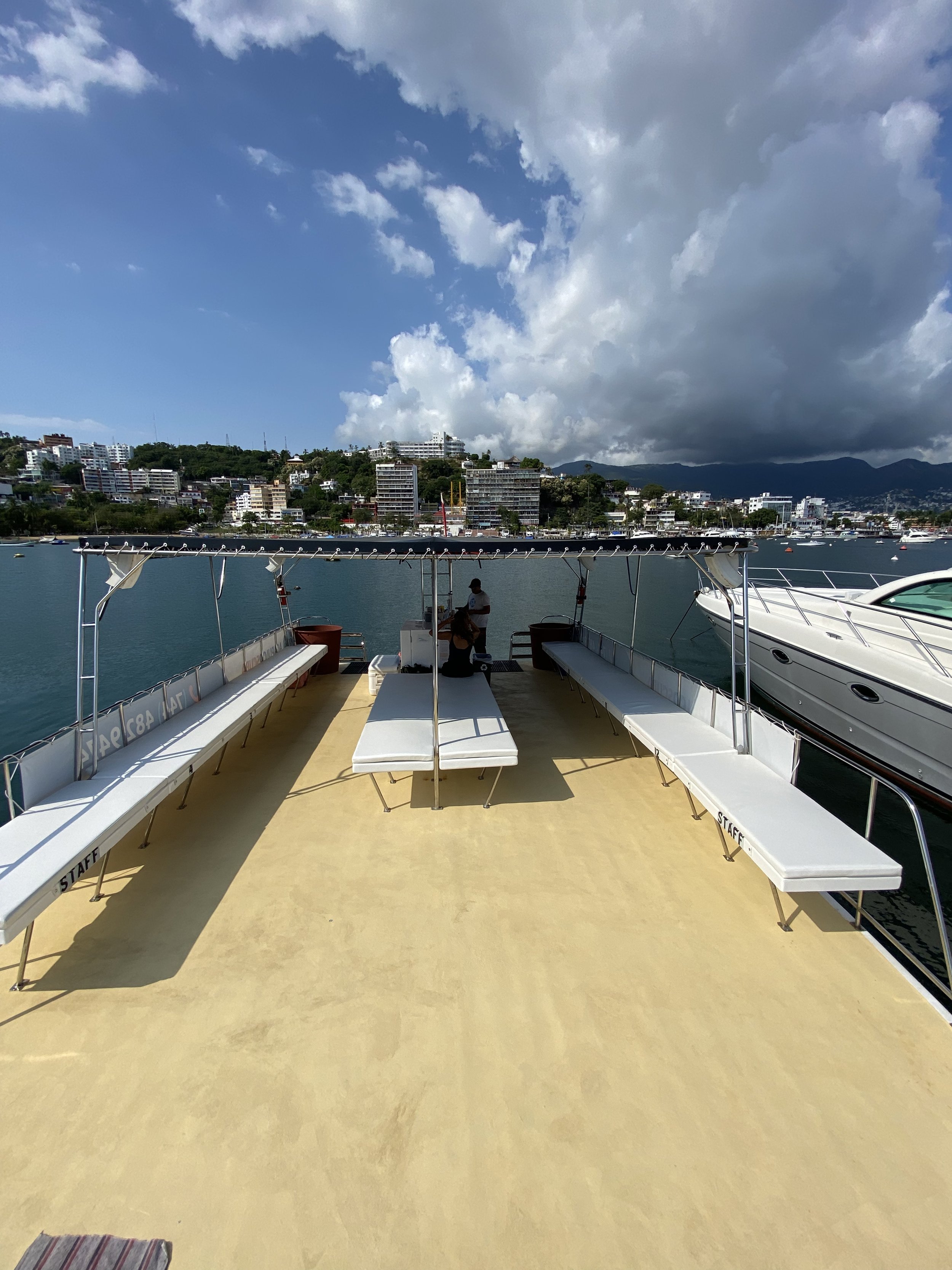 View from the deck of a boat, with seating benches on each side and a person sitting in the middle, overlooking a marina with boats and a cityscape with mountains in the background.
