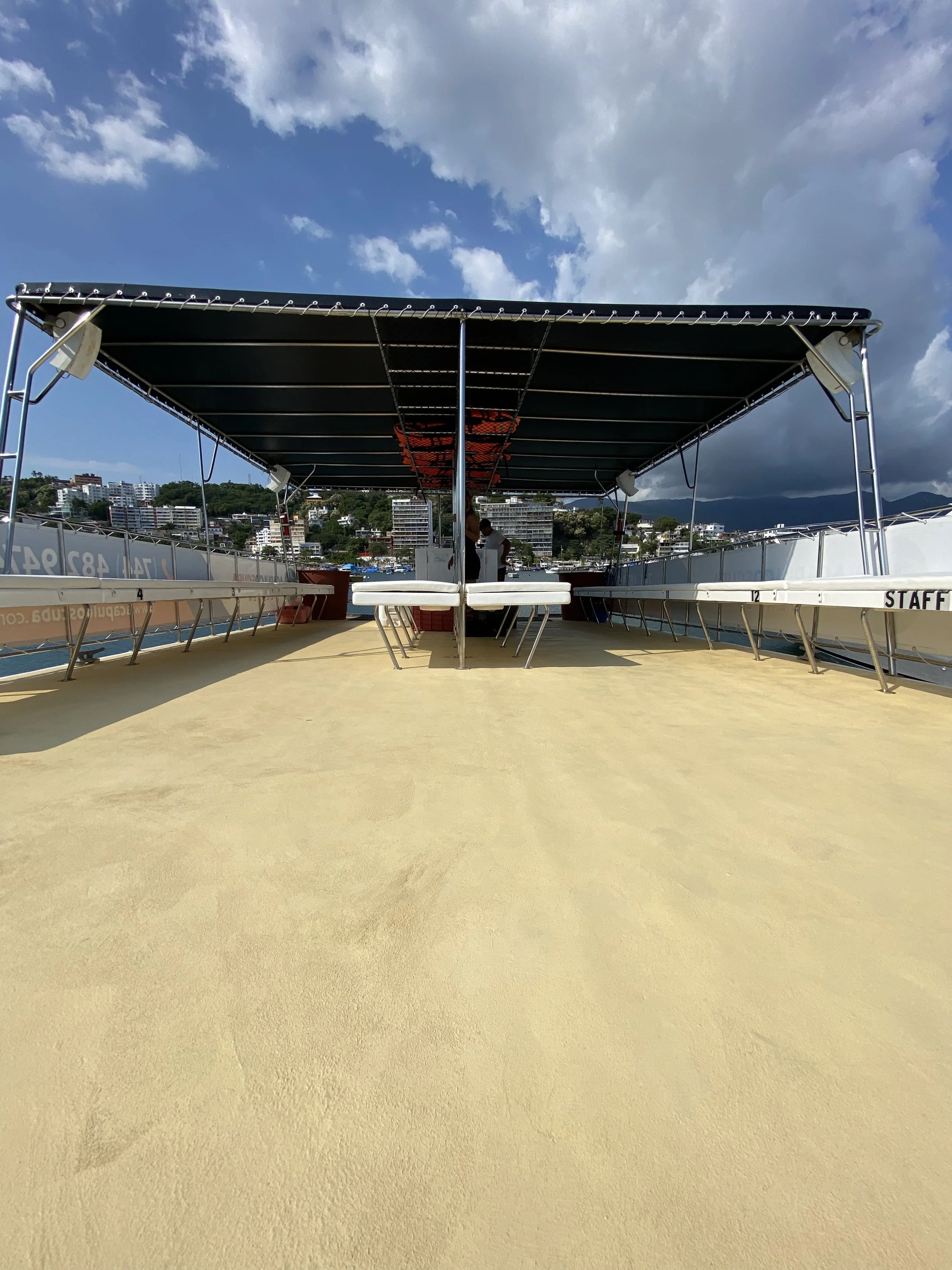 View of a boat deck with a canopy, seating, and a person standing at the back, overlooking a city with buildings and hills under a partly cloudy sky.
