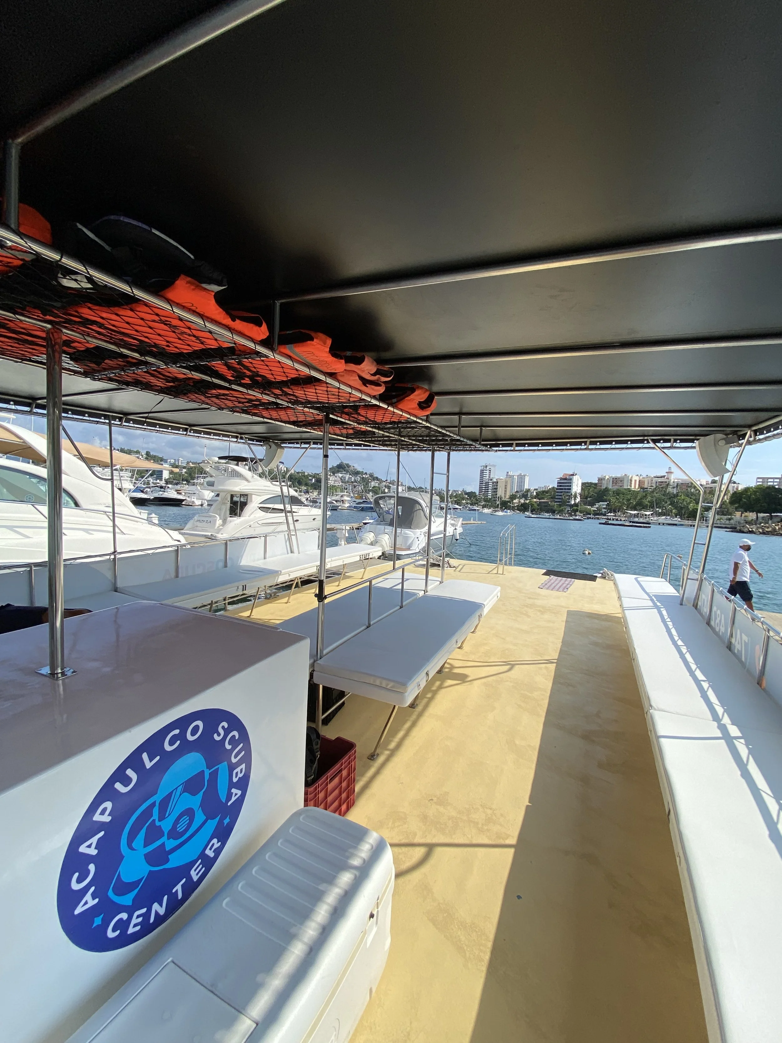 View of a boat deck with life jackets, benches, and a cooler, overlooking a marina with yachts and a city skyline in the background.