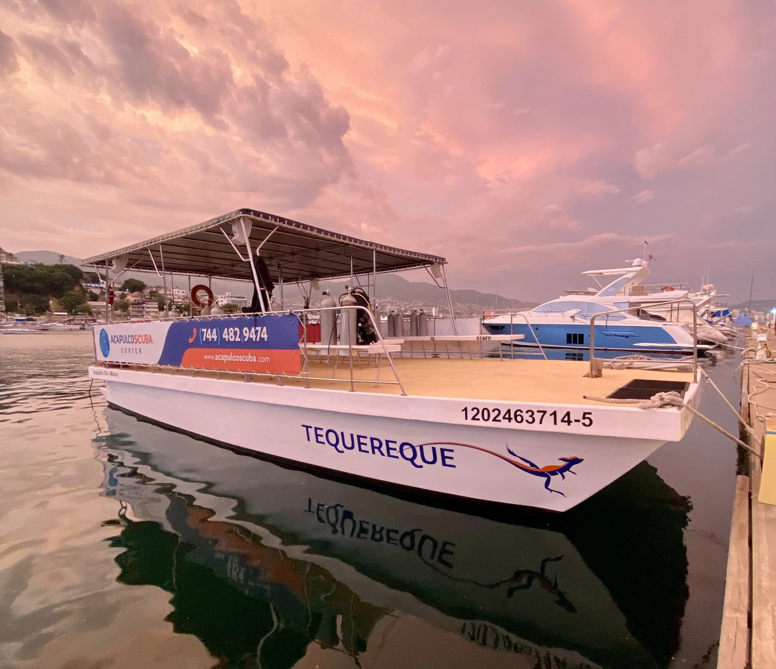 A boat docked at a marina during sunset with a pink sky, calm water, and other boats in the background.