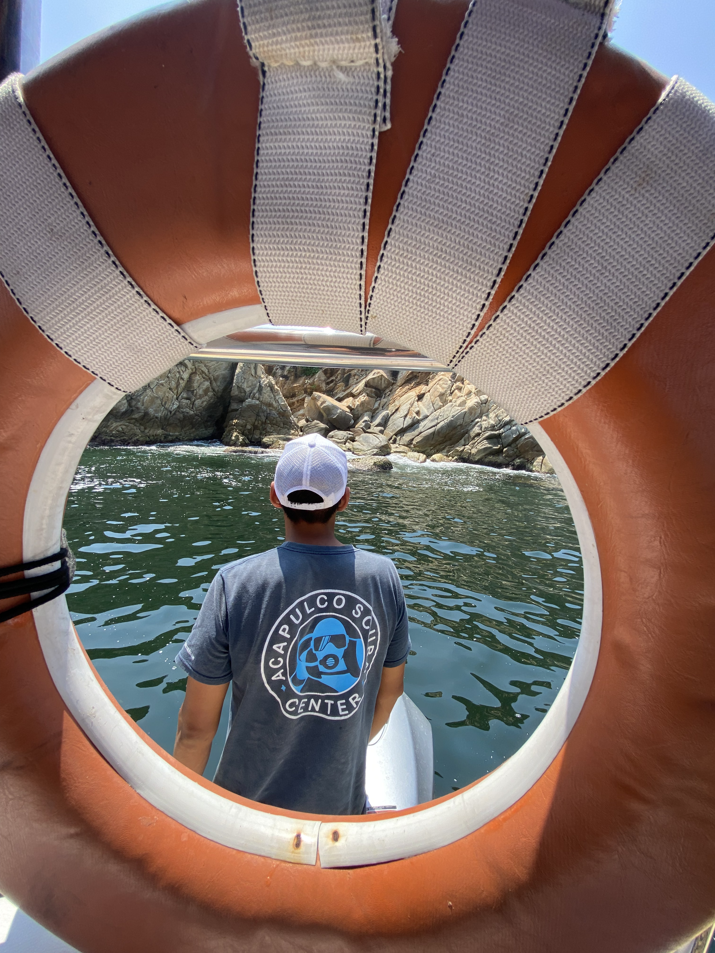 Person wearing a blue shirt and white cap seen from behind, sitting in a boat on water, framed by an orange and white life preserver.