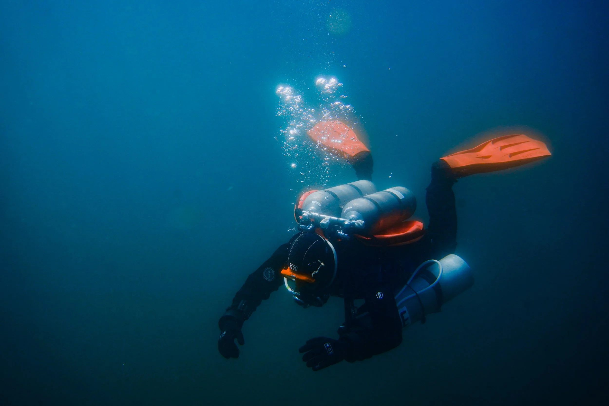 Scuba diver underwater wearing black wetsuit, orange fins, and carrying scuba tank and equipment, swimming in deep blue water.