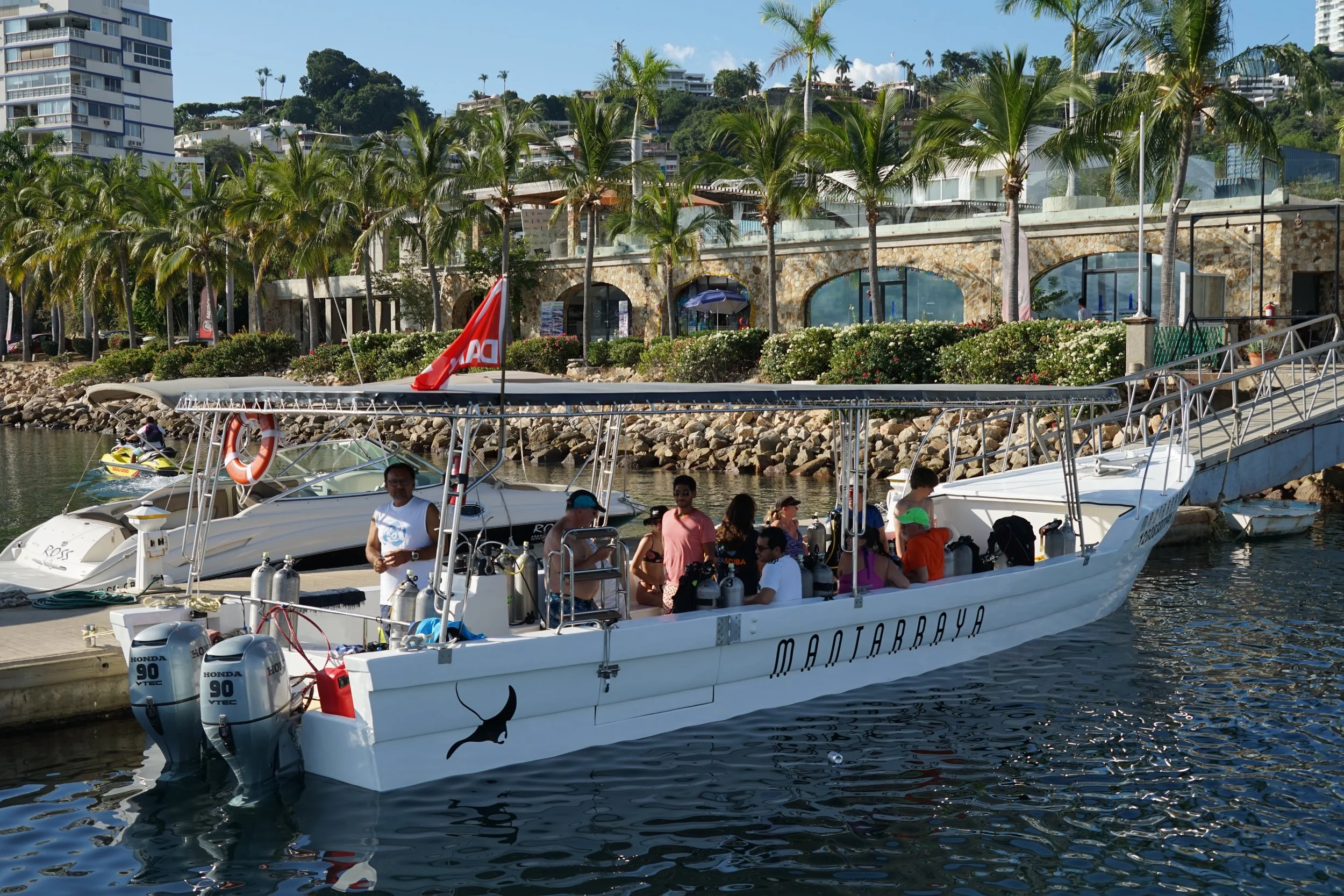 People on a white boat named 'Madrarraya' docked at a marina with palm trees and buildings in the background.