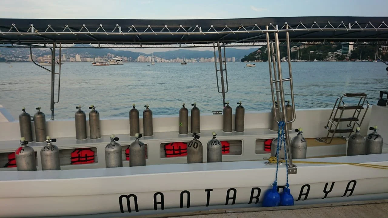A white boat equipped with multiple scuba tanks lined up along the side, set against a harbor with several boats and a city skyline in the background.