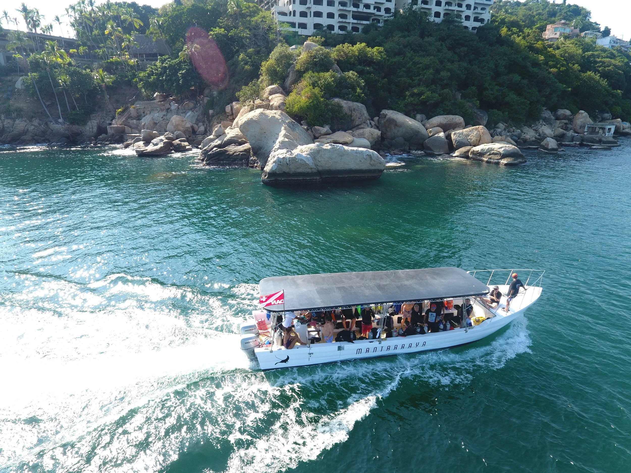 A white boat with a canopy carrying multiple passengers sailing near rocky, tree-covered coastline with large boulders and a hillside featuring residential buildings.