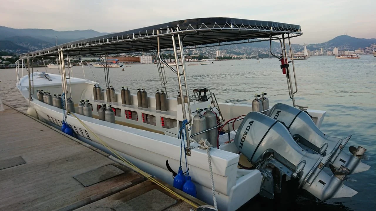 Passenger boat with multiple oxygen tanks on deck, docked at a pier with scenic city and mountain backdrop.