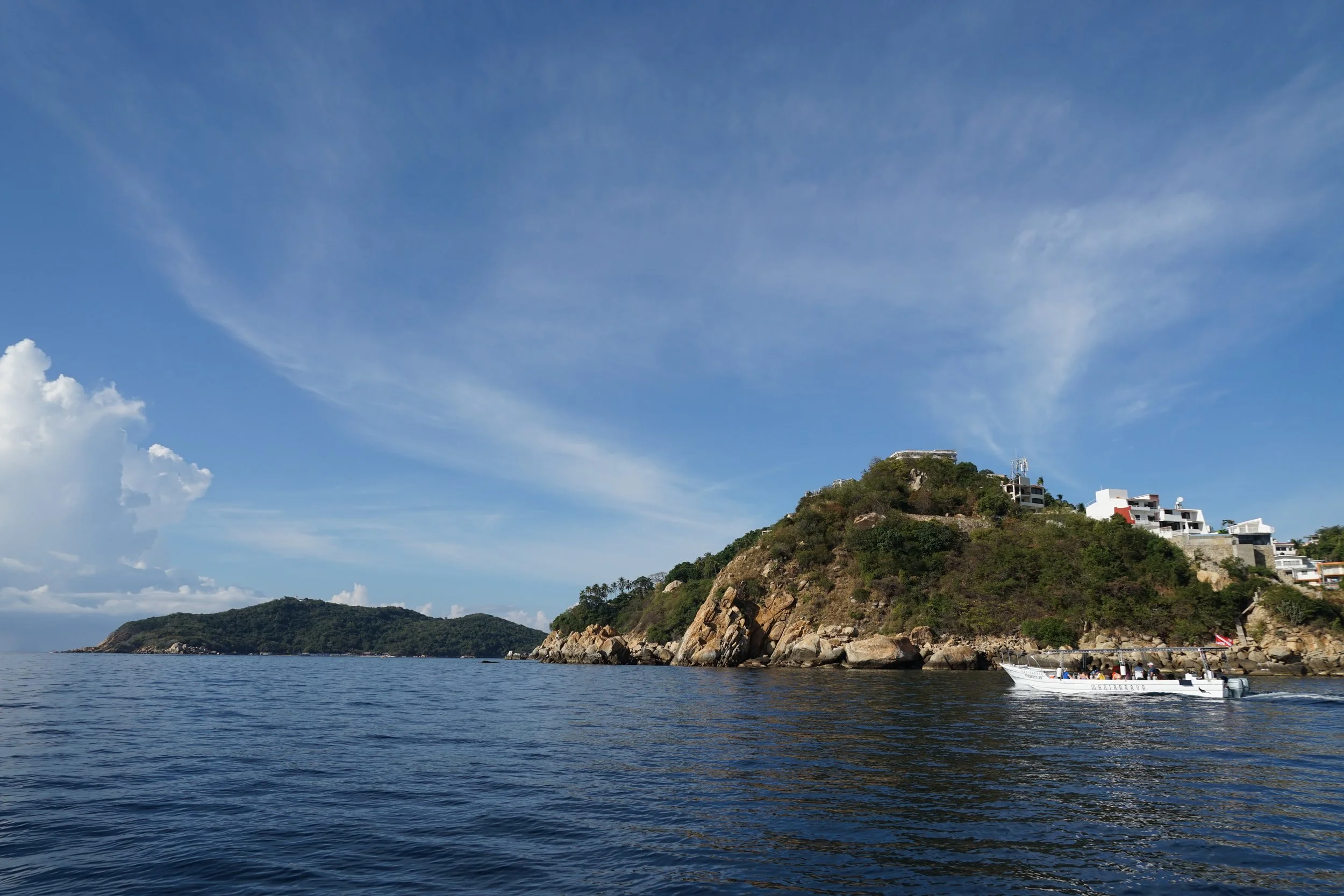 A boat on calm ocean waters near a rocky hillside with green vegetation and modern white houses, under a partly cloudy blue sky.