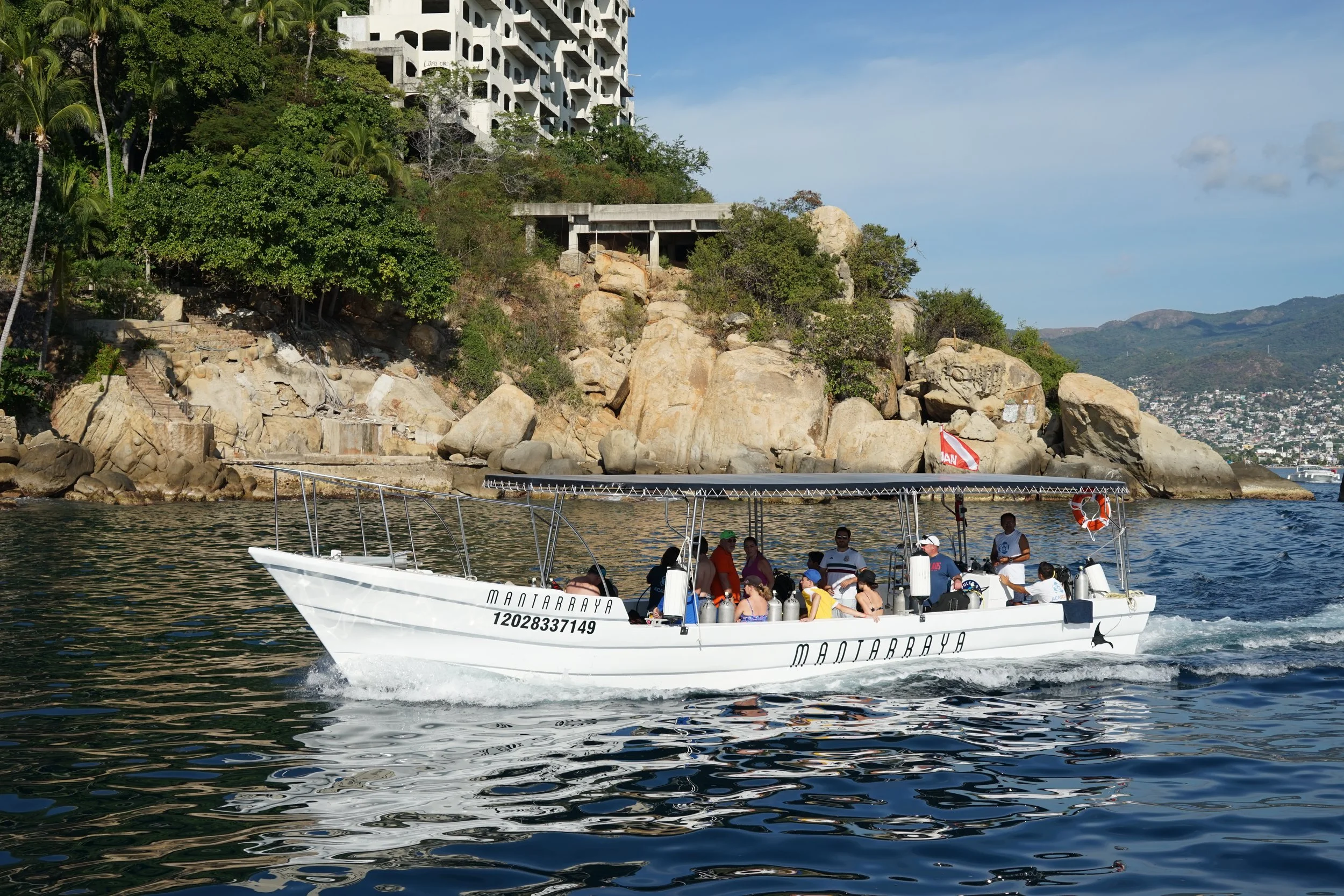 A group of people on a white boat named MANTARRAYA sailing on calm water near a rocky coastline with greenery and large buildings in the background.