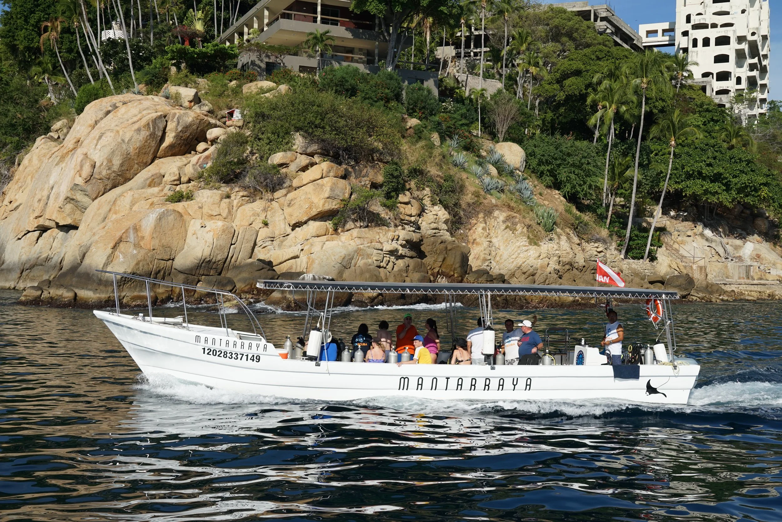 A boat named 'MANTARRAYA' with people onboard, sailing near a rocky coastline with buildings and palm trees in the background.