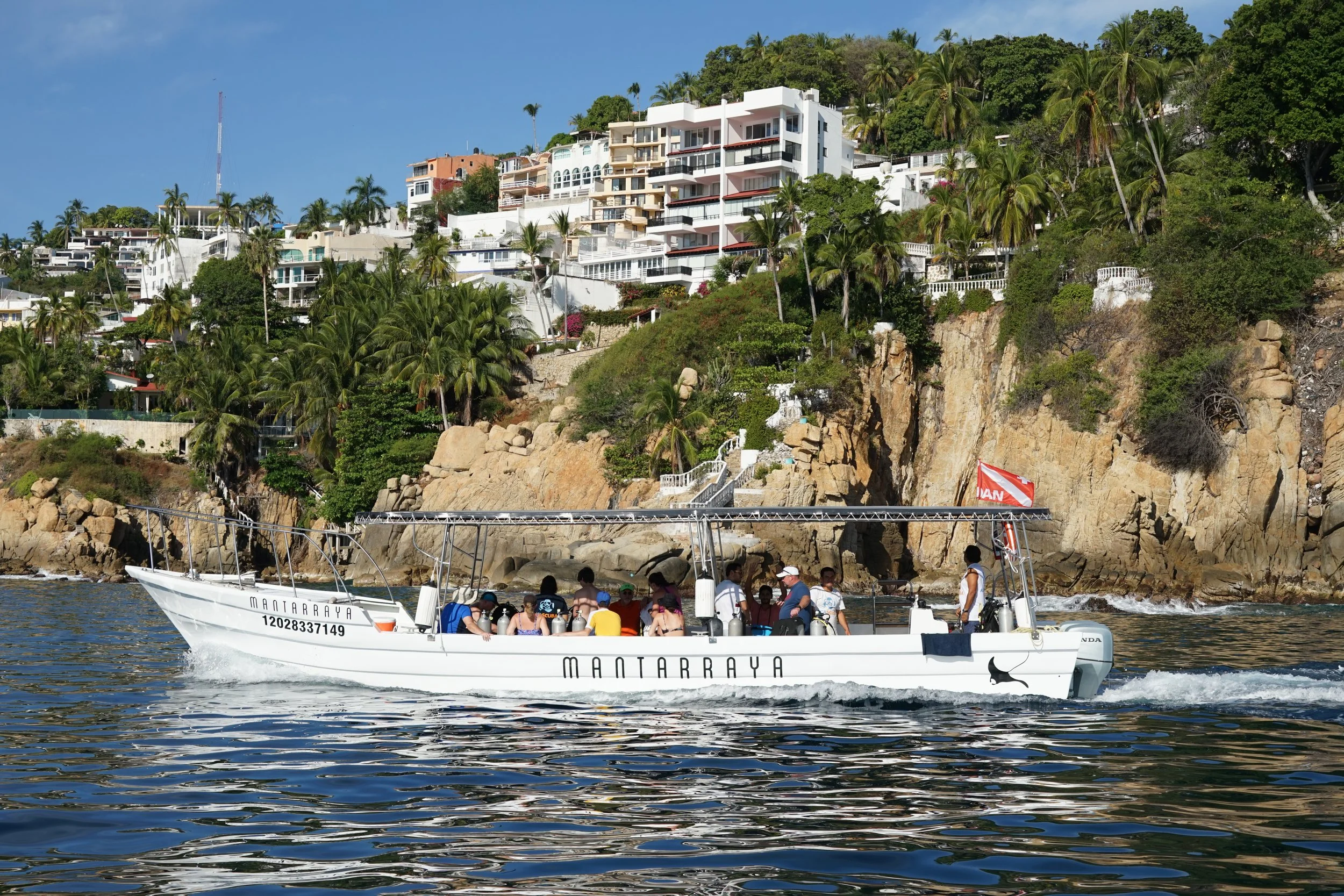A white boat labeled 'MANTARRAYA' with people on board, floating on water in front of a hillside with houses and palm trees.