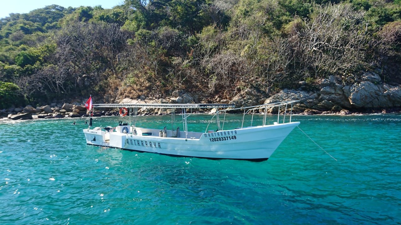 White boat named 'Autarraiya' anchored near rocky shoreline with green trees, on clear blue water.