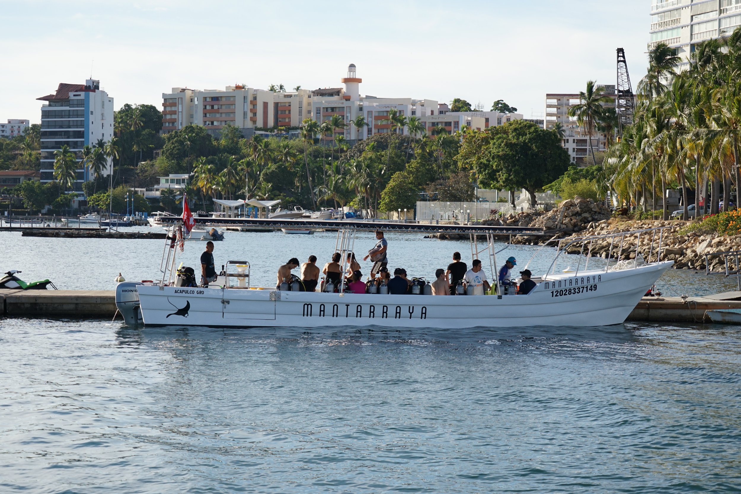 A group of people on a white boat named Mantarraya docked in a marina with palm trees, buildings, and a rocky shoreline in the background.