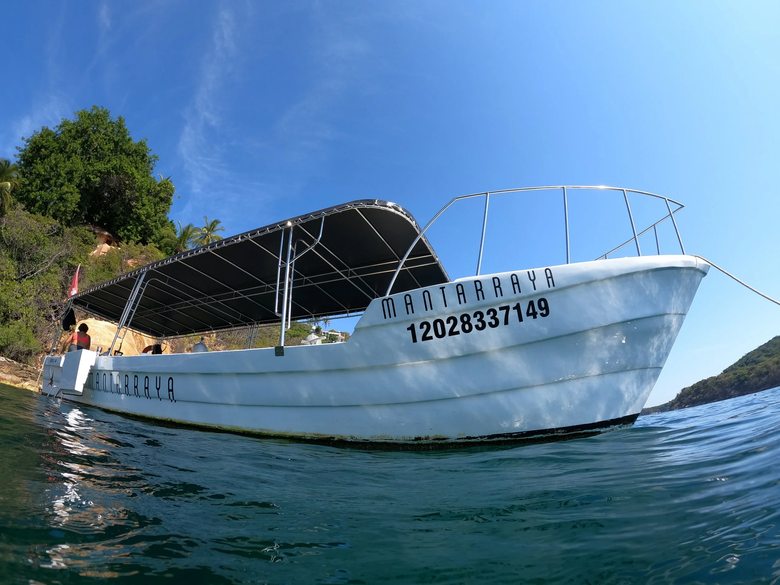 A white boat named MANTARRAYA floating on water near a rocky shoreline with green trees, under a clear blue sky.