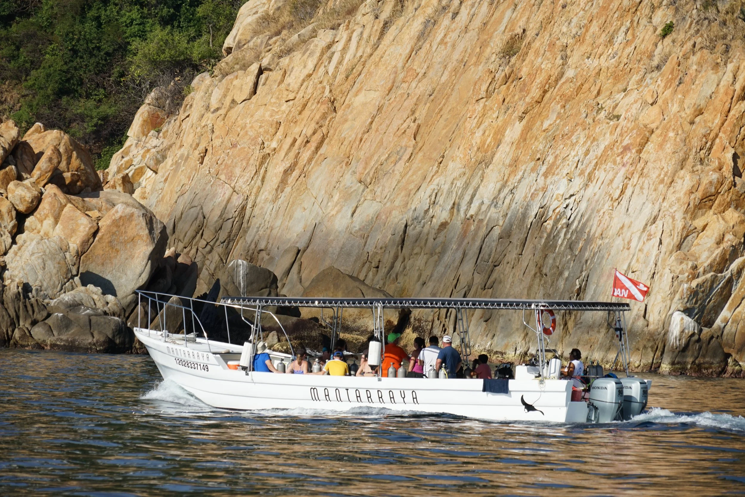 A white boat named 'MARTARRAYA' with several people on board, sailing near a rocky cliff, with calm water and a few trees in the background.