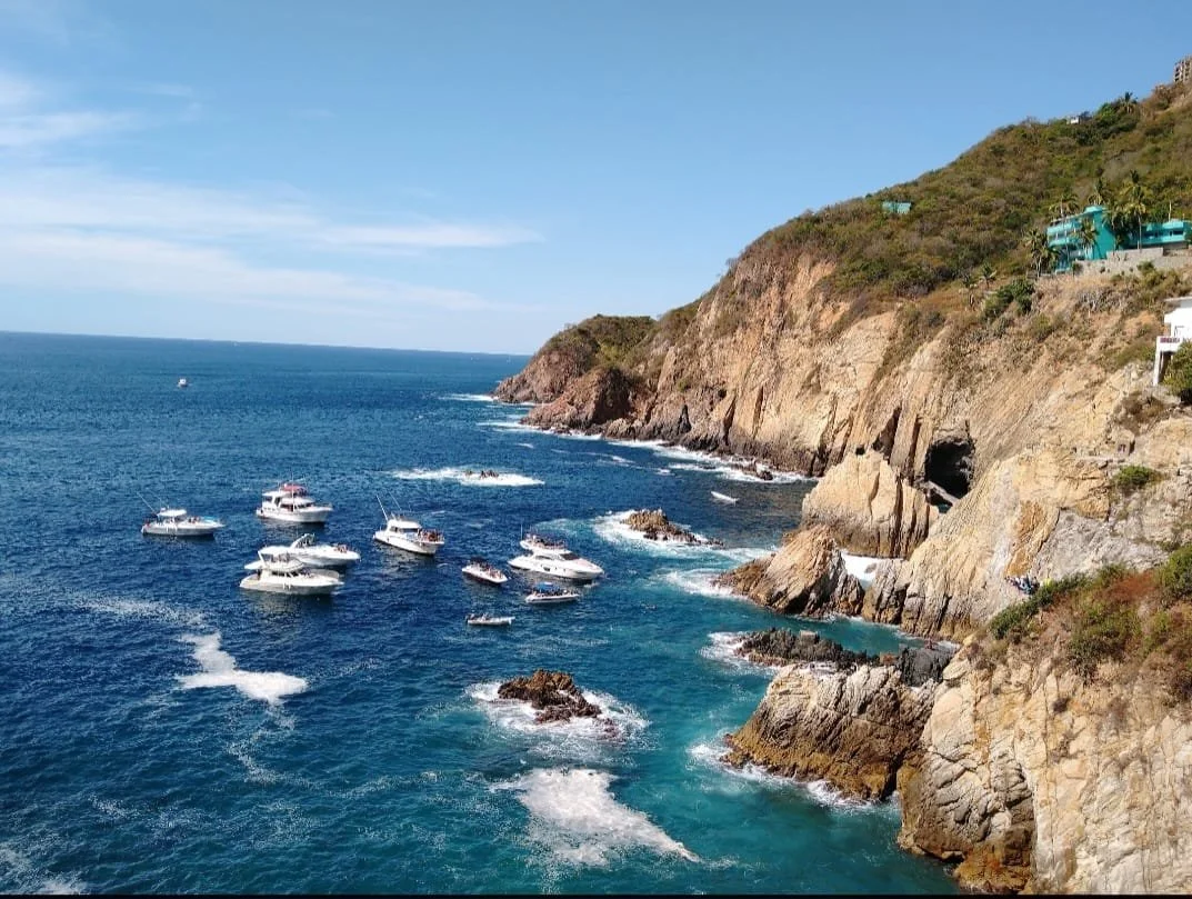 Boats anchored near a rocky coastline with cliffs and buildings on hilltop, under a blue sky.