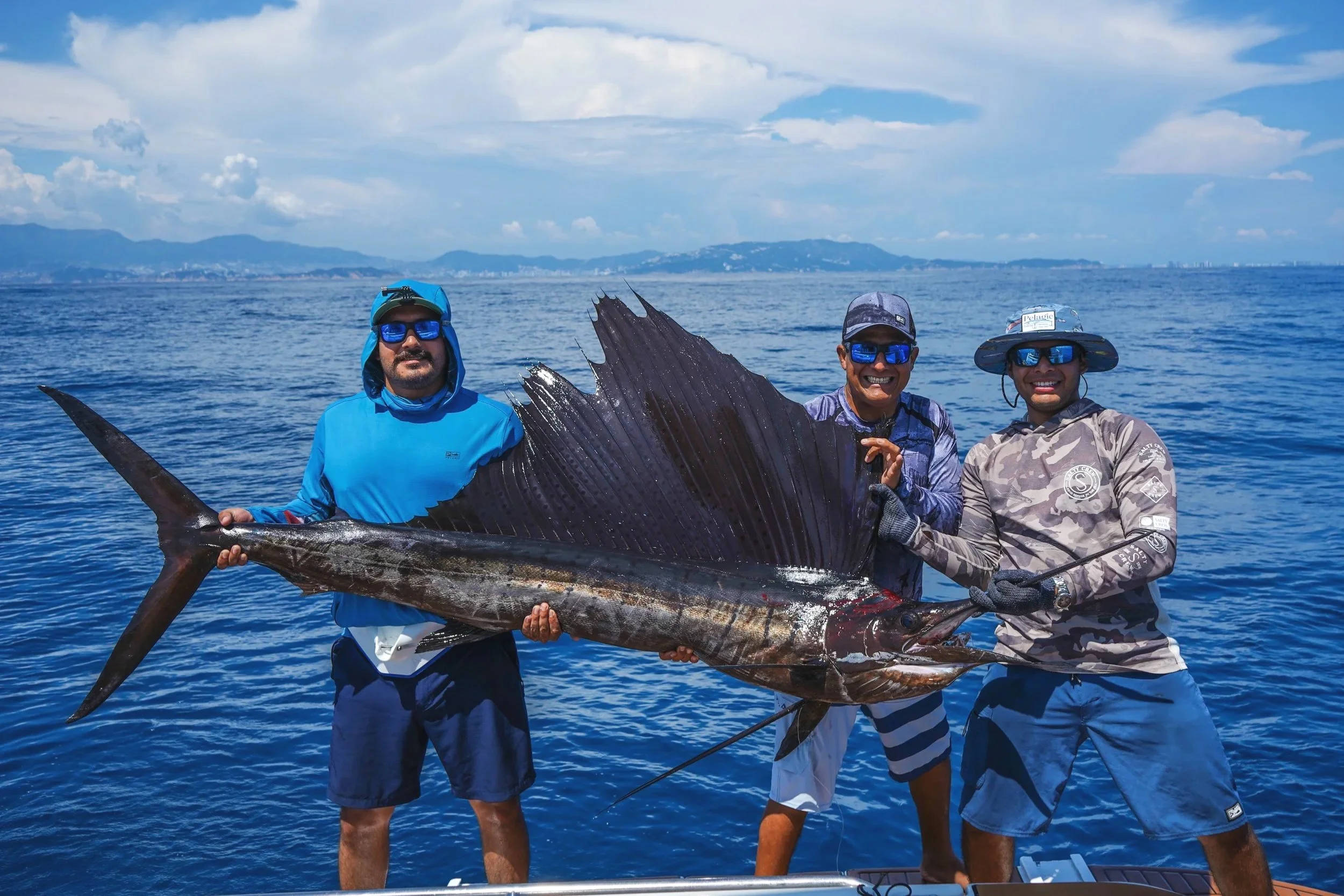 Three men on a boat holding a large sailfish with a scenic ocean and distant mountains in the background