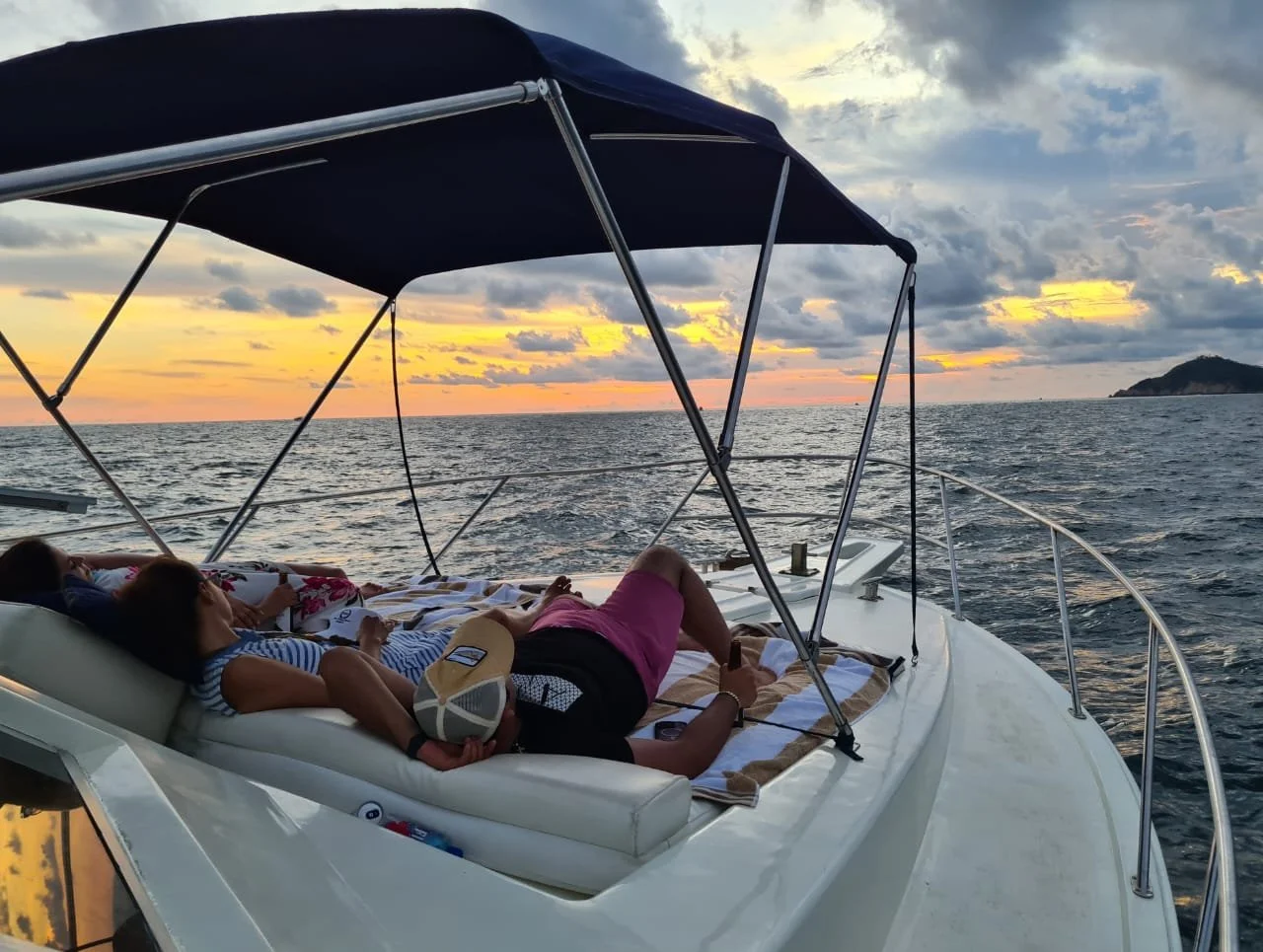 People relaxing on a yacht at sunset, with the ocean and a distant island in the background.