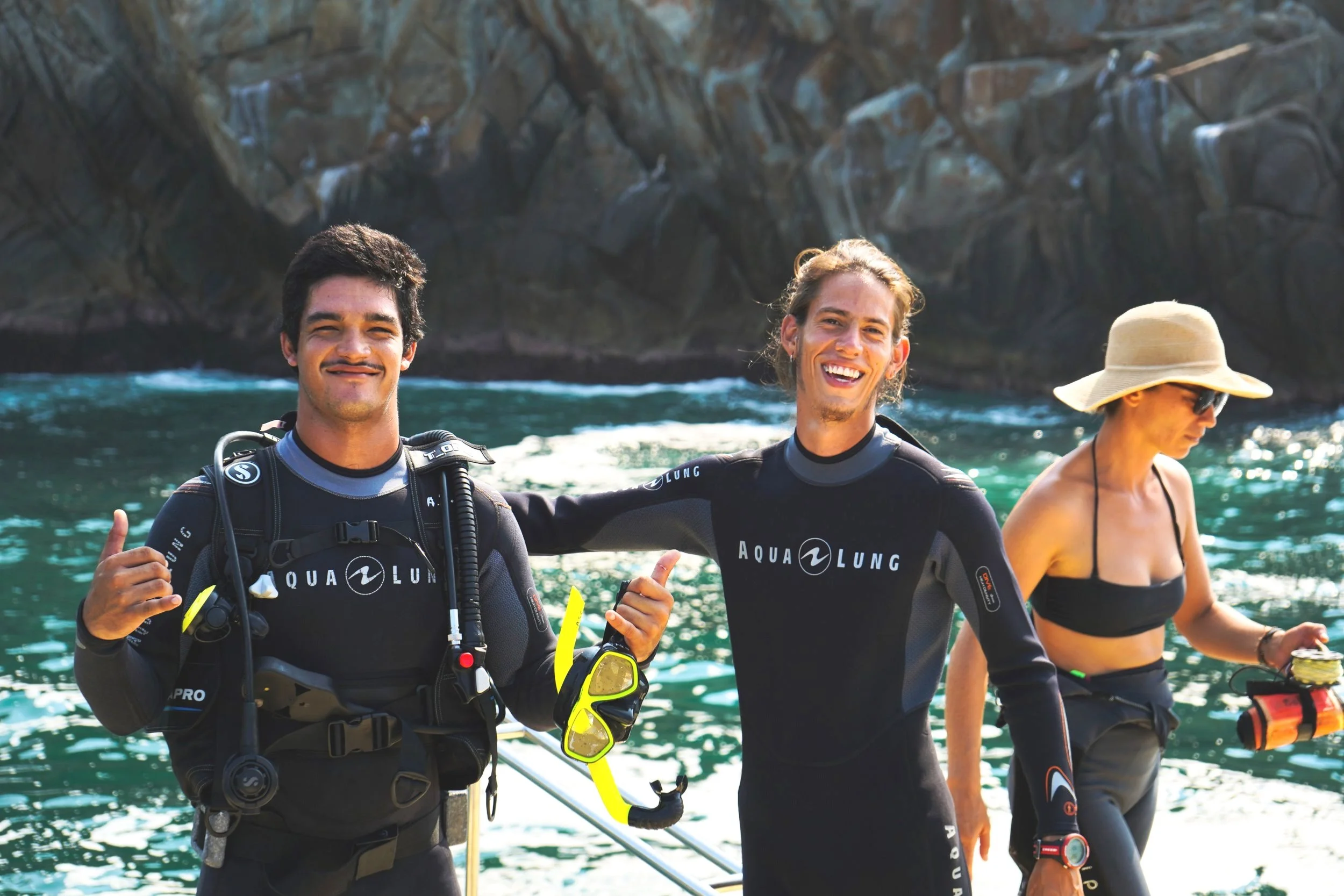 Two people in wetsuits smiling and showing a thumbs-up pose in front of water, with a woman in the background holding diving gear and wearing sunglasses and a hat.