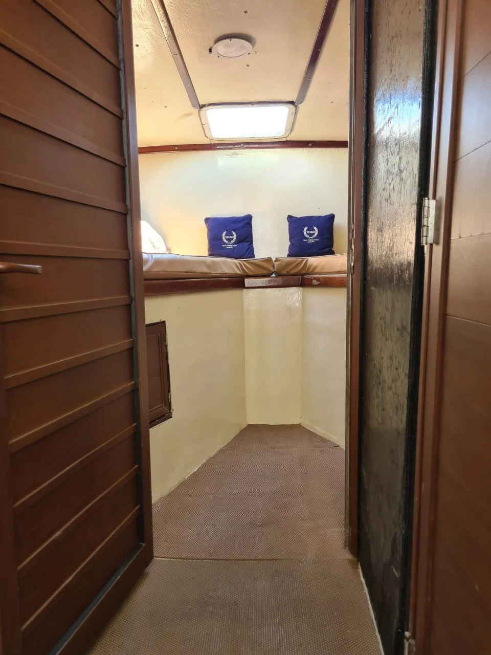 Interior of a small boat cabin with a view into the sleeping area containing a low bed with two blue pillows, a small window on the ceiling, and wooden walls.