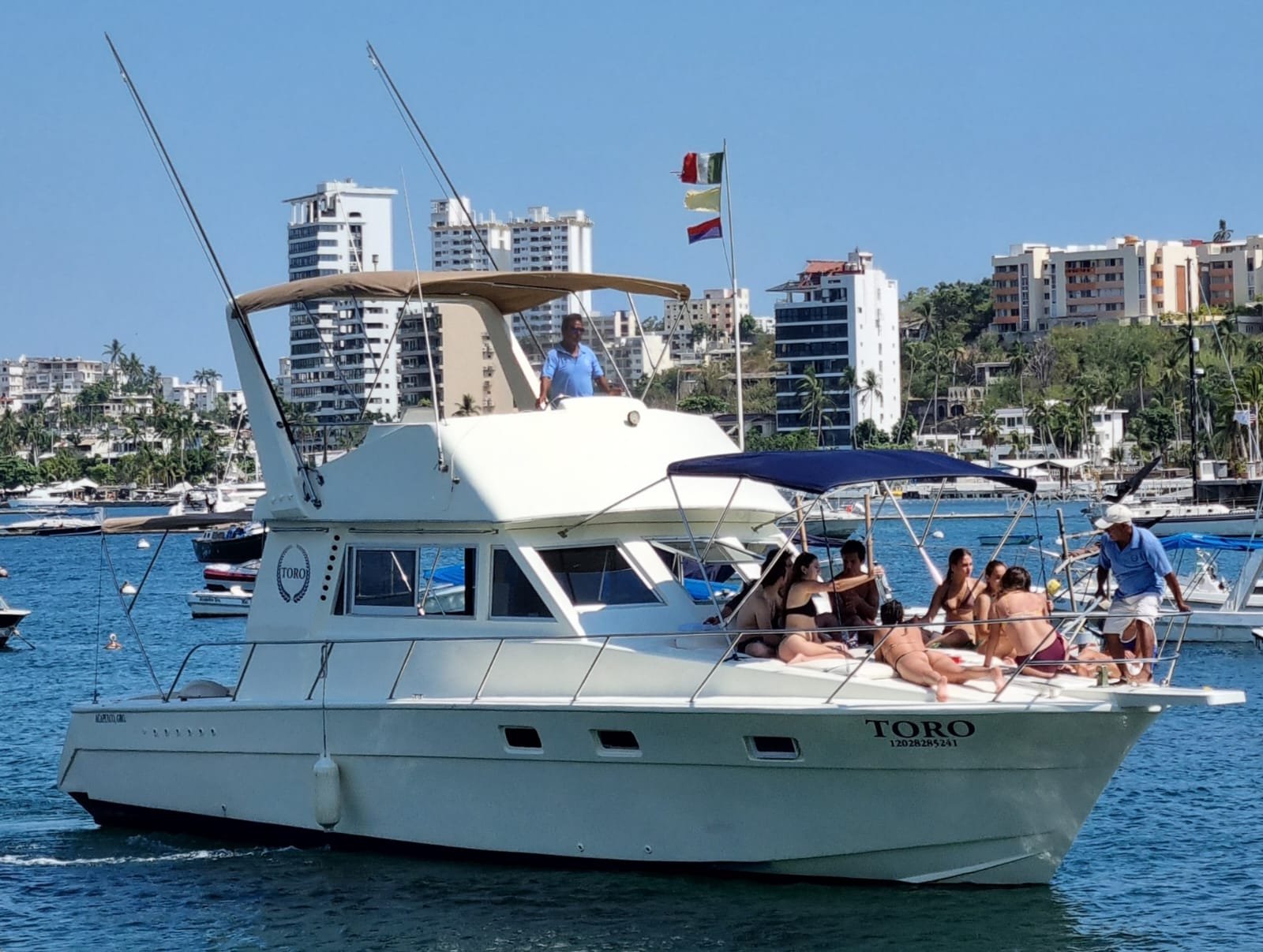A white yacht named TORO cruising in a harbor with people sunbathing and socializing on the deck, and a city skyline with high-rise buildings and greenery in the background.