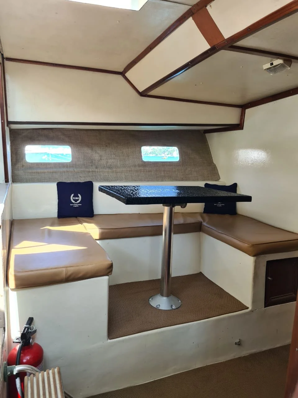 Interior view of a boat cabin with a built-in L-shaped beige cushioned bench, a black table with a metal support in front, small rectangular windows, and a fire extinguisher near the entrance.