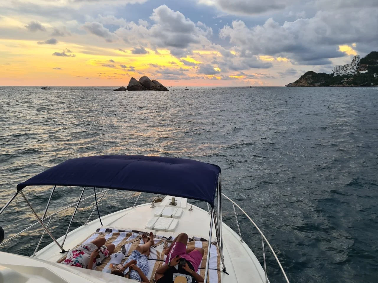 View from a boat on the water during sunset with cloudy sky, rocky island in the distance, and a boat with children lying on deck under a blue canopy.