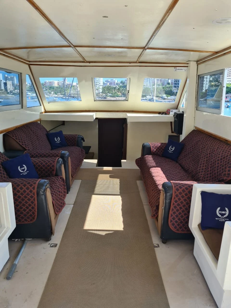 Interior of a boat with two maroon-patterned couches, each with a navy blue pillow, facing each other in a small lounge area. There are large windows showing a marina and cityscape outside. The ceiling is beige with wooden trim, and there is a centra