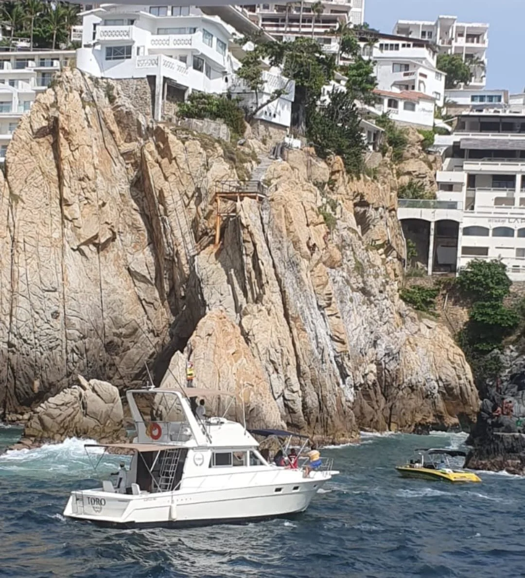 A white motor yacht sailing near a rocky cliff with luxurious white houses on top, overlooking the ocean.