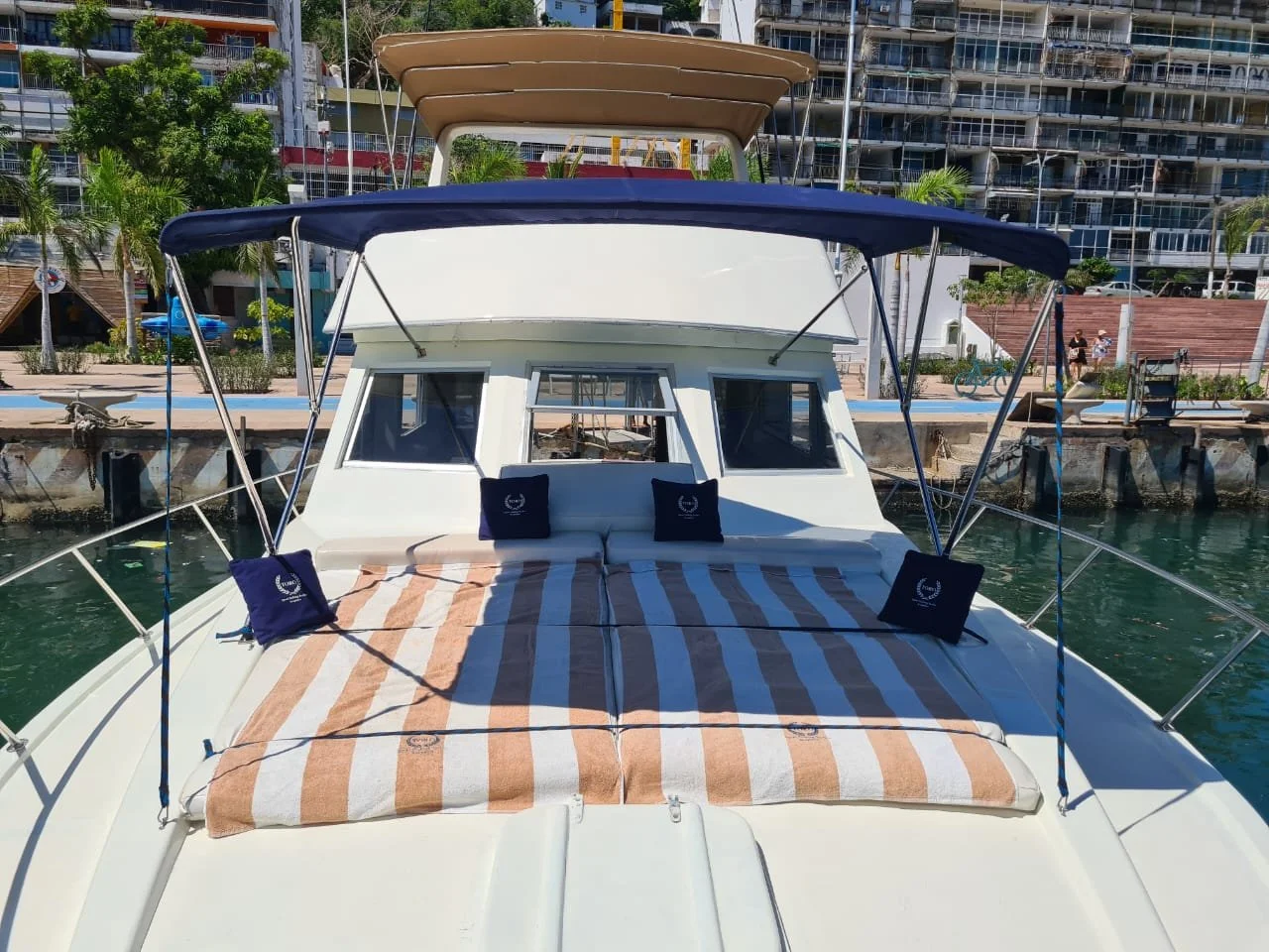 Front view of a white yacht docked at a marina with a striped towel on the bow, blue and beige sunshades, and city buildings in the background.