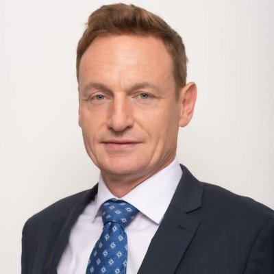 Professional headshot of a man in a dark suit, white shirt, and blue patterned tie, against a plain light background.