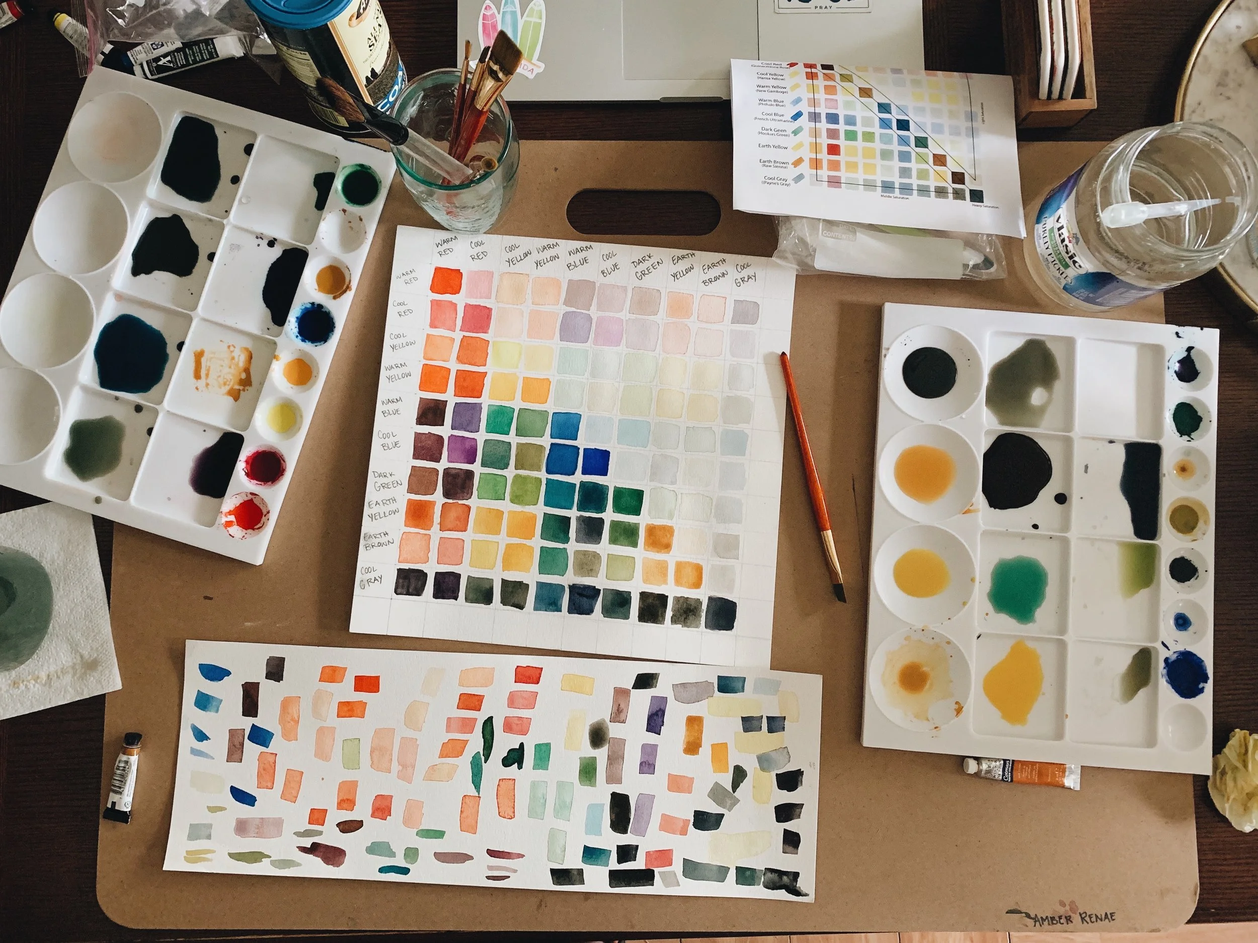 A watercolor painting workspace with color charts, paint palettes, brushes, a jar of water, and color reference sheets on a table.