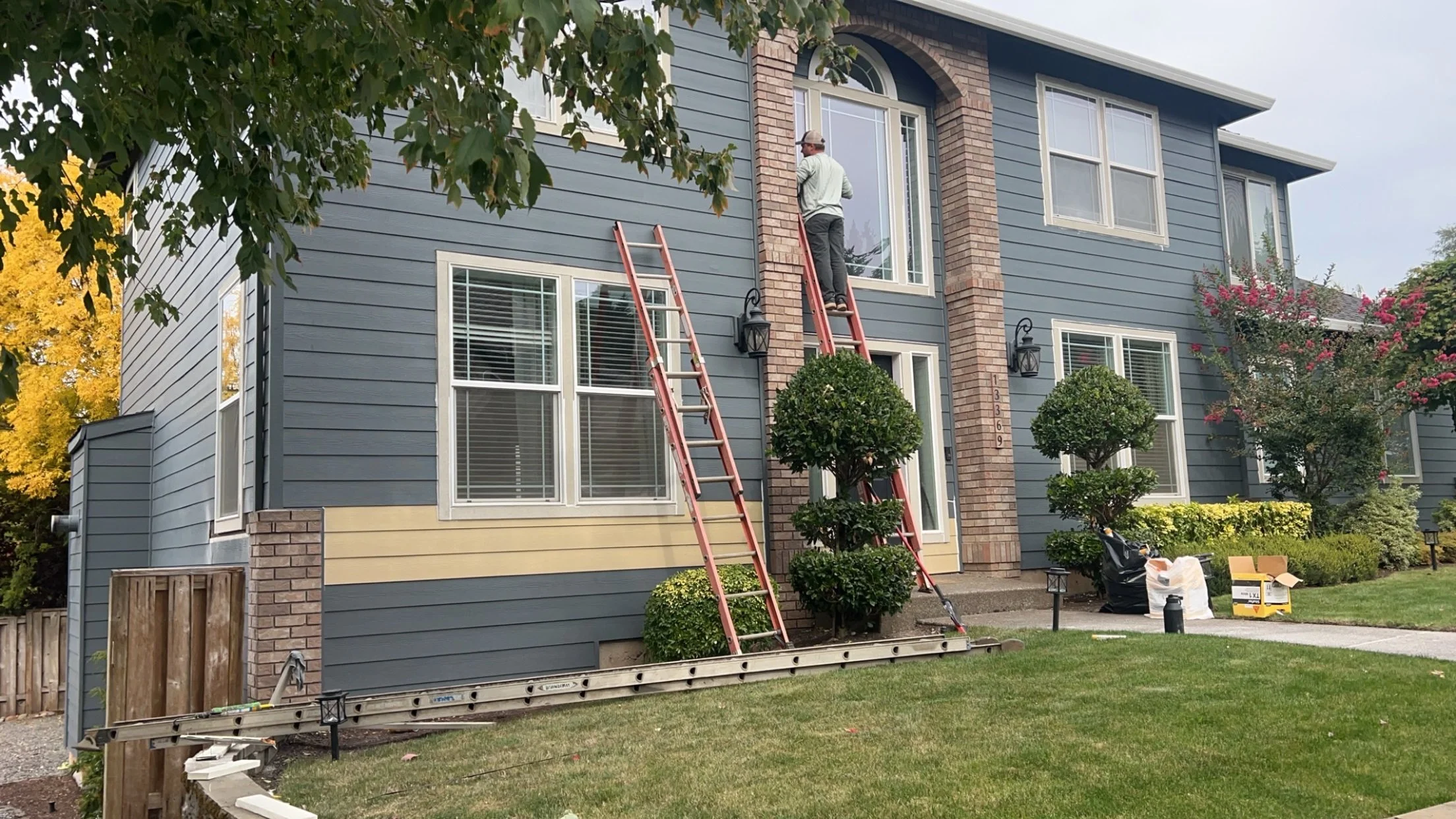 A house renovation scene with a man on the second-floor window installing or working on the window, two ladders leaning against the house, and gardening shrubs in front. The house has blue and yellow siding with brick accents, and the yard is landscaped with grass, trees, and shrubs.