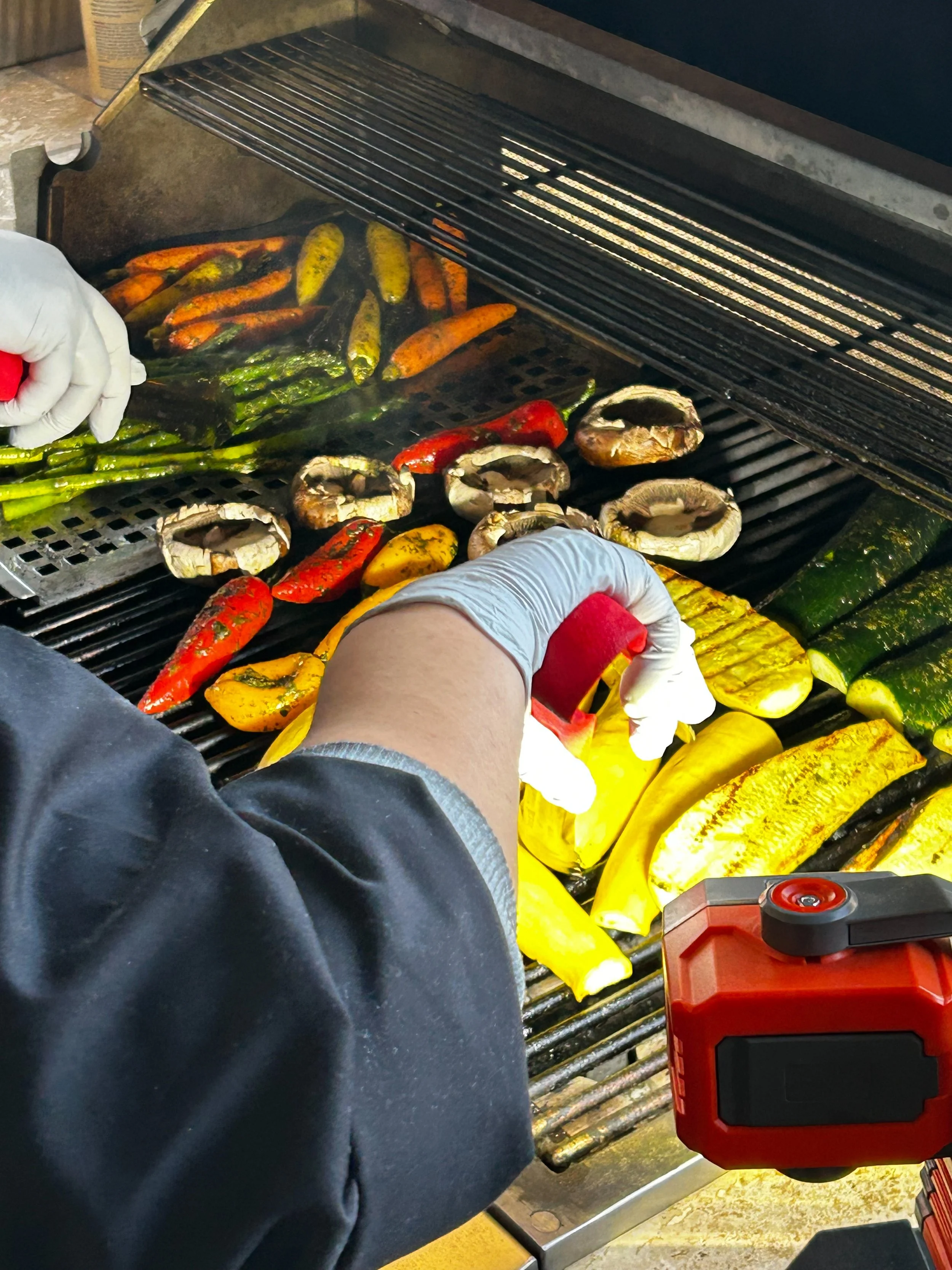 Person grilling colorful vegetables, including red and yellow peppers, zucchini, and mushrooms, on a barbecue grill outdoors.