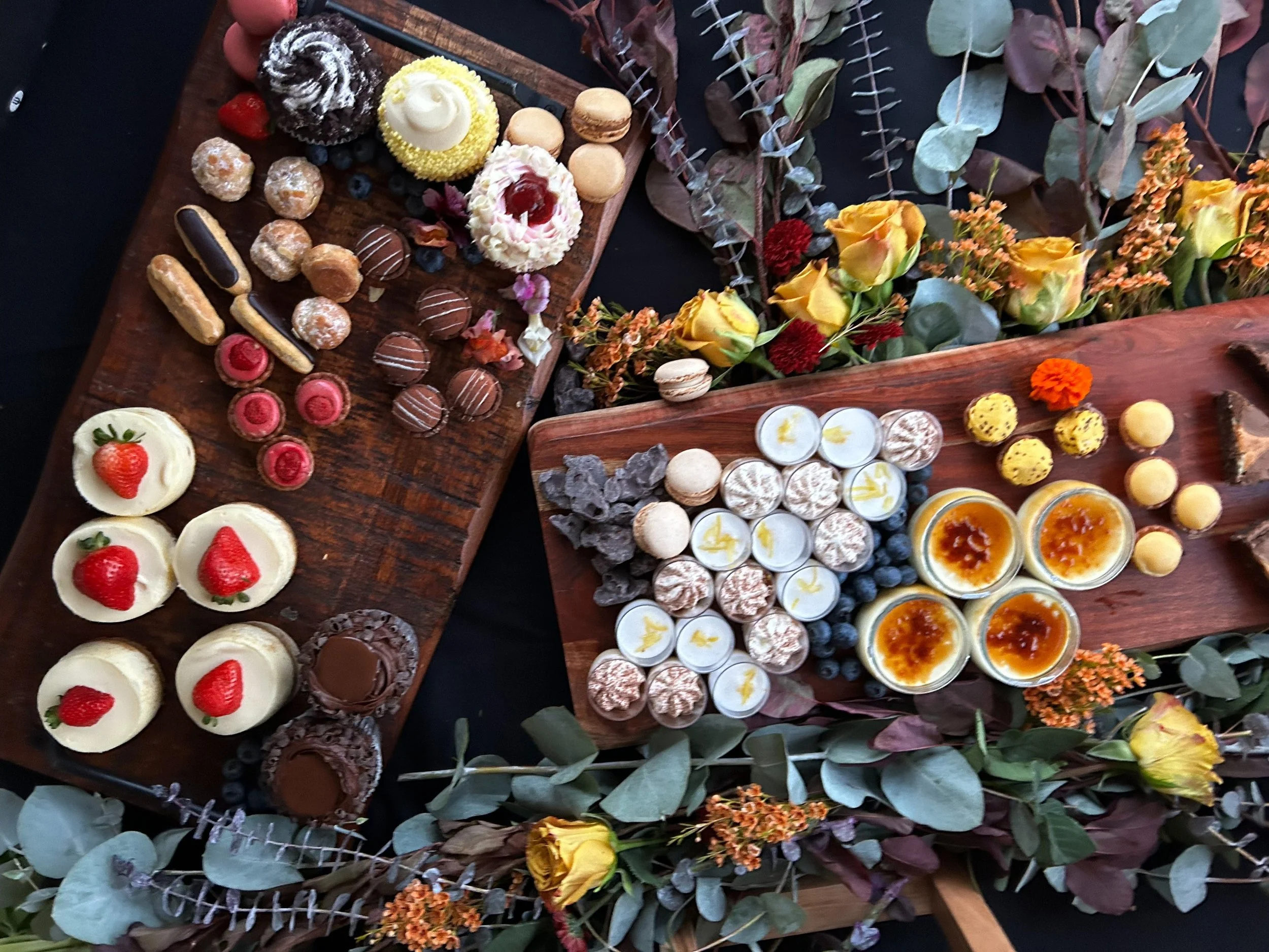 A top-down view of three wooden platters filled with various desserts and surrounded by floral decorations. The left platter has strawberry-topped cheesecakes, pink and chocolate truffles, éclairs, and mini cakes. The middle platter features white an