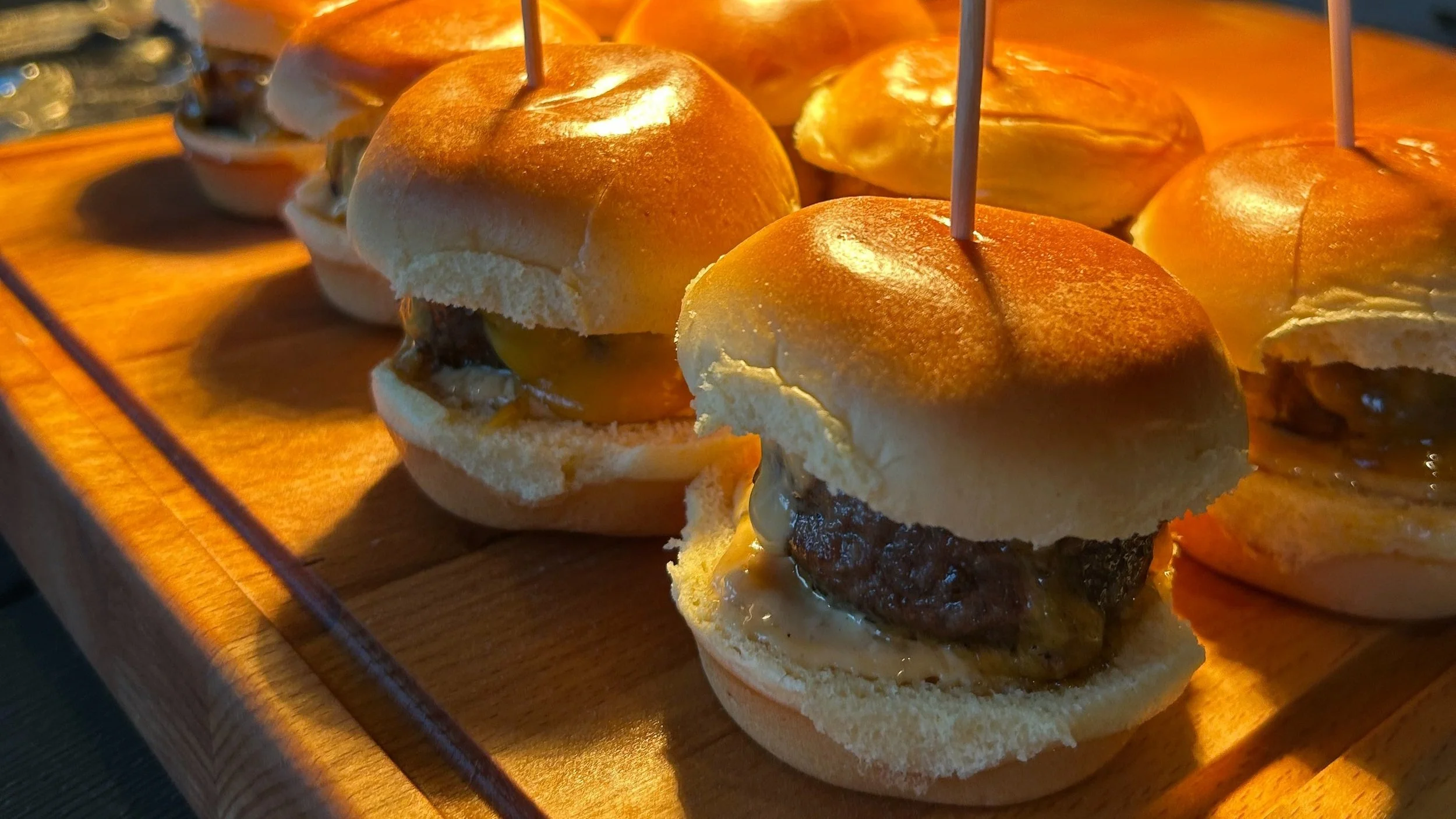 Close-up of mini cheeseburgers topped with small sandwich buns and skewered on toothpicks on a wooden serving board.